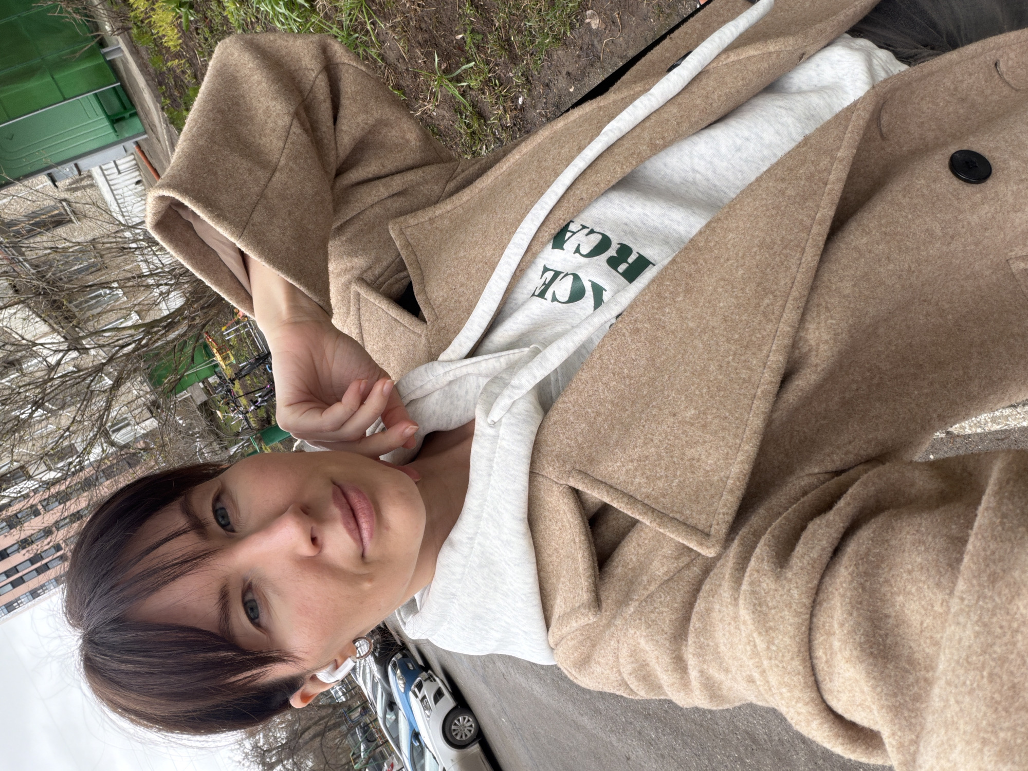 The image shows a selfie taken by a woman with short brown hair and blue eyes, who is wearing a beige coat and a light gray hoodie. She is wearing a white wireless earbud in her left ear. The woman is holding the camera at arm's length, capturing her face and upper body.
The background reveals a street scene, with parked cars visible on the left side of the frame. The area appears to be urban, with trees and buildings visible in the background. The environment suggests a residential or commercial district with pedestrian access. Based on the visible surroundings, the location is identified as the city of Perm, Russia.