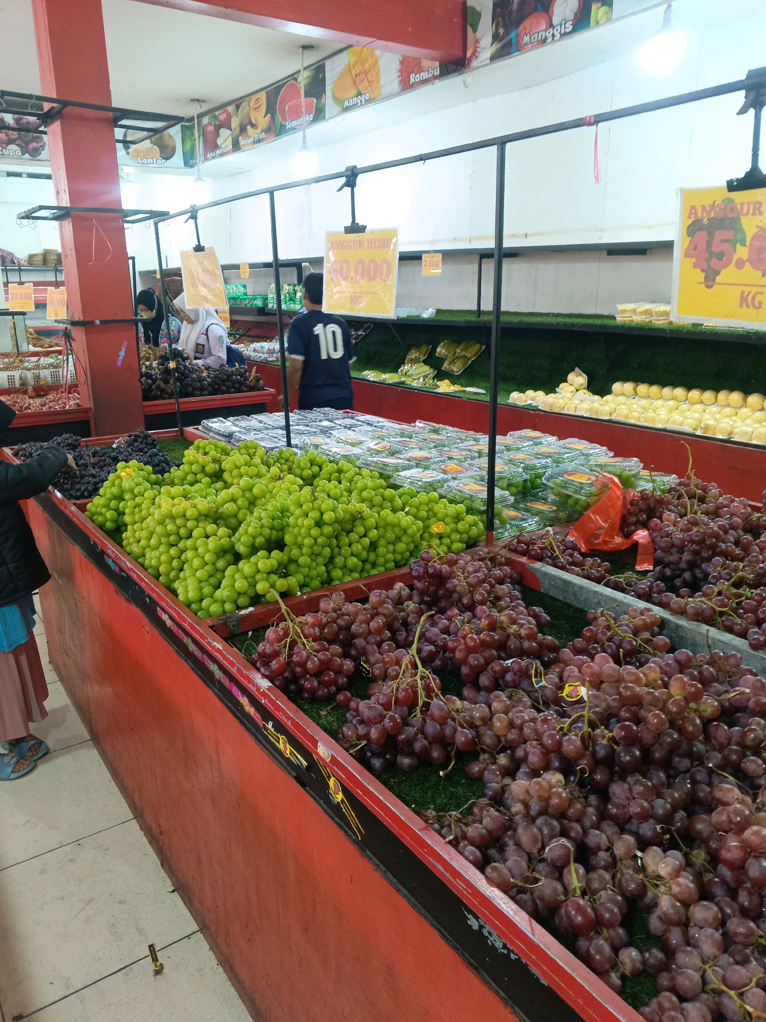 The image depicts a fruit market stall, likely located in the city of Garut, Indonesia, given the presence of signage with prices in Indonesian Rupiah (e.g., "60.000"). In the foreground, an assortment of red and green grapes is displayed in large bins. Several bunches of red grapes are prominently featured, with some appearing to be slightly overripe. To the left, a child wearing a pink skirt and blue sandals is reaching into a bin of dark-colored grapes.
Behind the grapes, there are rows of packaged fruits, possibly grapes or small apples, in clear plastic containers. Further back, a vendor wearing a black shirt with the number "10" on it is visible. To the left of this vendor, two individuals wearing white and dark headscarves are interacting with the fruit display. Above the stall, various fruit images are displayed on posters, including mangosteen, rambutan, and what appear to be mangoes and watermelon. The overall environment suggests an indoor market setting with artificial lighting. The flooring appears to be tiled.