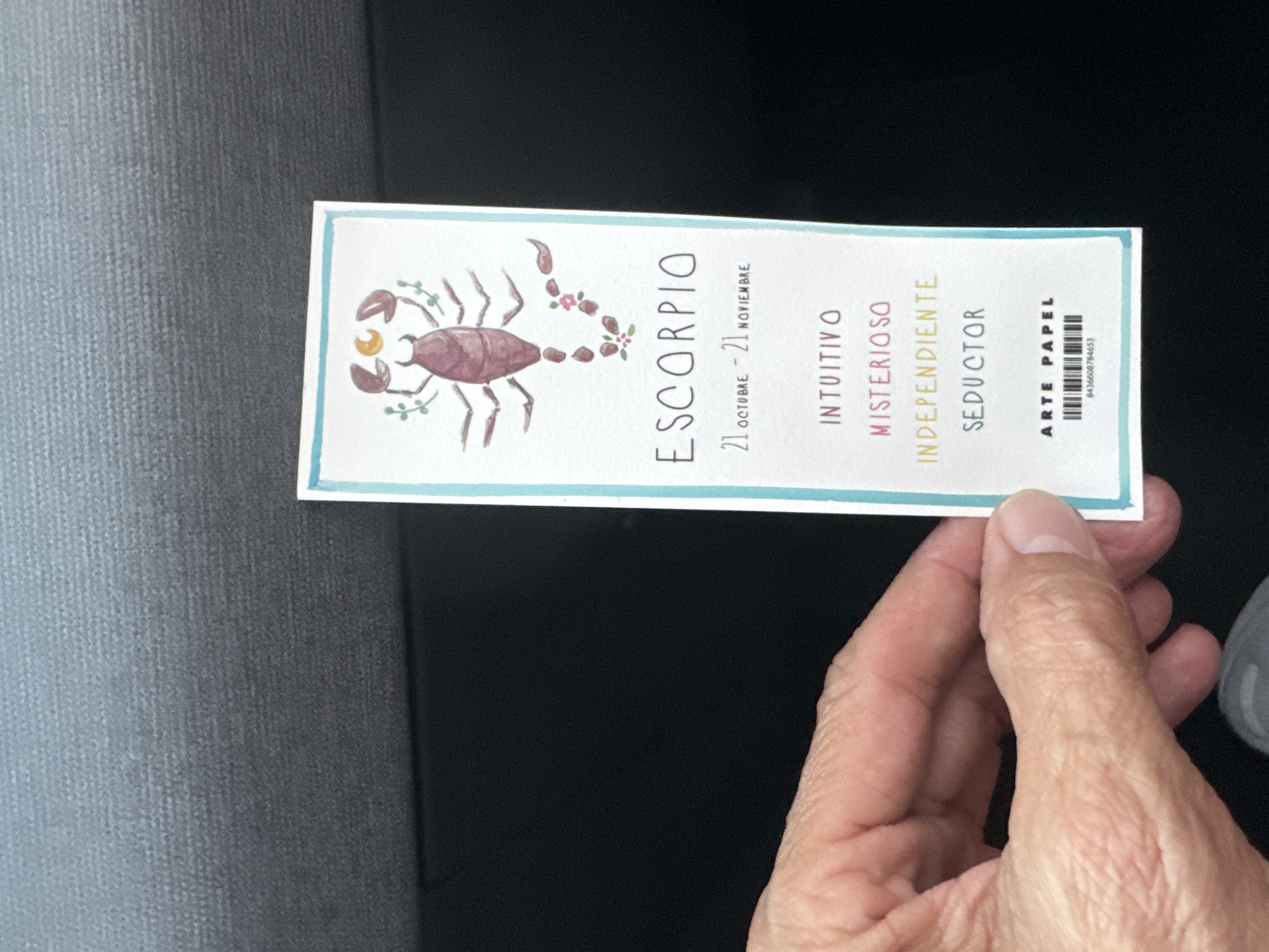 The image shows a person's hand holding a rectangular bookmark with a watercolor illustration of a scorpion at the top. Below the scorpion, the text reads "ESCORPIO" in large letters, followed by the dates "21 OCTUBRE - 21 NOVIEMBRE." Further down, there are four descriptive words in different colors: "INTUITIVO," "MISTERIOSO," "INDEPENDIENTE," and "SEDUCTOR." At the bottom of the bookmark, it says "ARTE PAPEL" and a barcode with the number "8436608784653." The bookmark has a light blue border. The background of the image is out of focus and appears to be a textured gray surface, possibly upholstery, and a dark area below. There is no discernible information within the image that links it to the city of Murcia, Spain.
