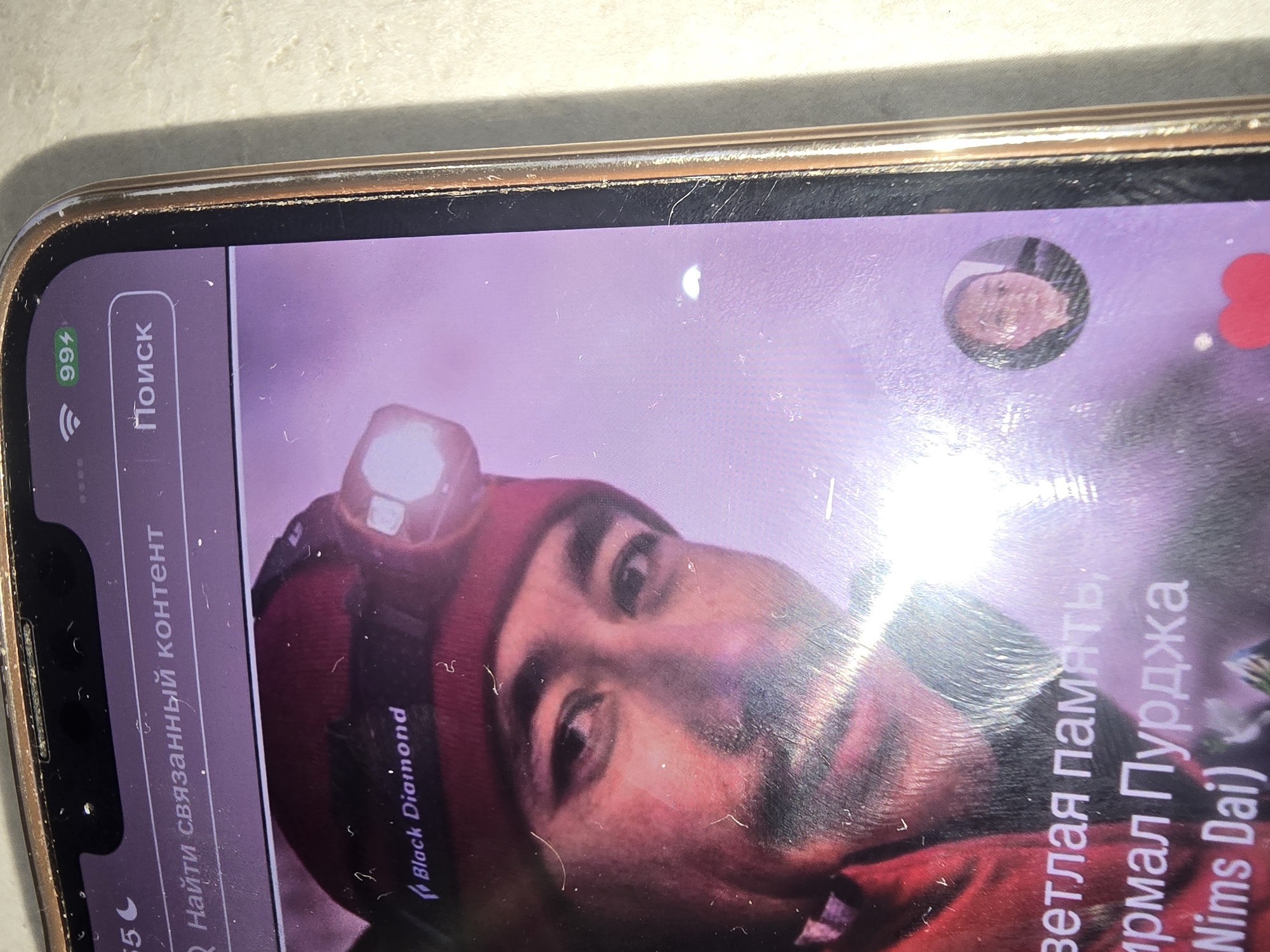 The image is a close-up of a smartphone screen displaying a video or image with text overlays. The screen shows a man wearing a red beanie with a headlamp. The text "Black Diamond" is visible on the beanie. Below the man's face, partially obscured text reads "ветлая память; ирмал Пурджжа (Nims Dai)". In the upper right corner of the screen, a battery indicator shows "99%". A small circular profile picture is visible in the lower right quadrant of the screen, displaying a young person's face. The phone itself is gold-rimmed and has some surface scratches. The background behind the phone is a light-colored, textured surface. The context of Ulan-Ude, Russia, cannot be determined from the visual information present in the image.