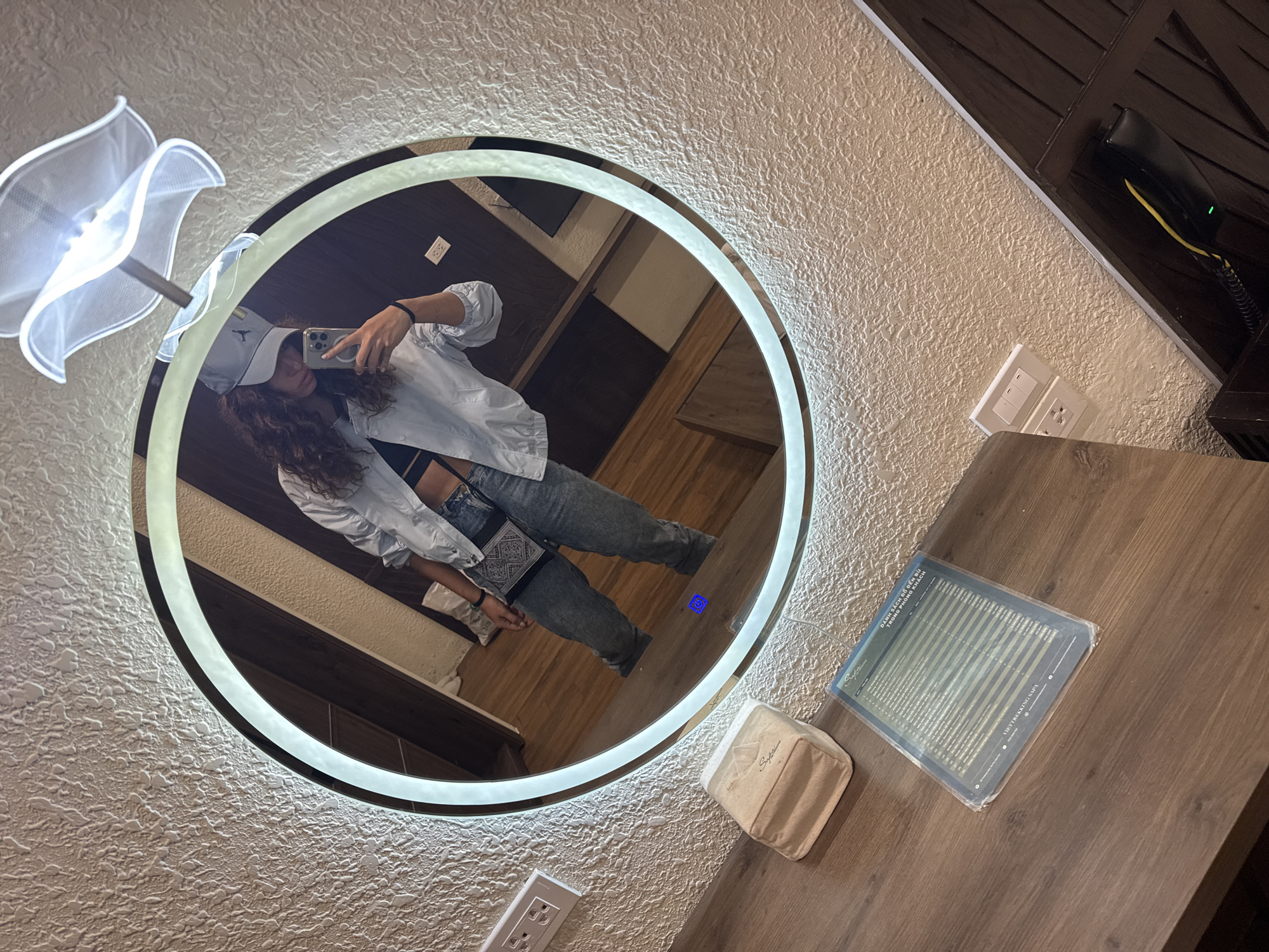A person is taking a selfie in a round, illuminated mirror. The person is wearing a white cap, a white shirt, jeans, and a black top. The mirror is located on a textured wall next to a wooden surface. On the wooden surface, there is a small white package and a document with a title that includes the words "SA PA" and "KHÁCH". The location is Sa Pa Ward, Vietnam. There is a lamp with white petals to the left of the mirror and a phone on a wall at the top right of the photo.