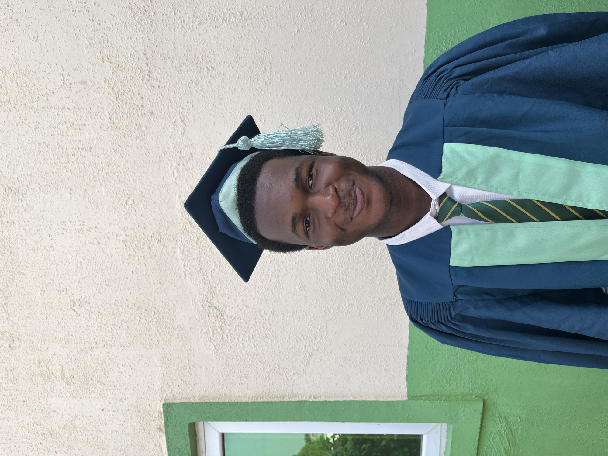 A young, dark-skinned man wearing a graduation gown and cap stands outdoors against a textured wall that is half cream and half green. The cap is dark blue with a light blue tassel. The gown is dark blue with a light blue stripe. He wears a white collared shirt and a green and gold striped tie. The bottom of a window frame with green trim is visible in the lower left corner. The image was taken in Jos, Nigeria.