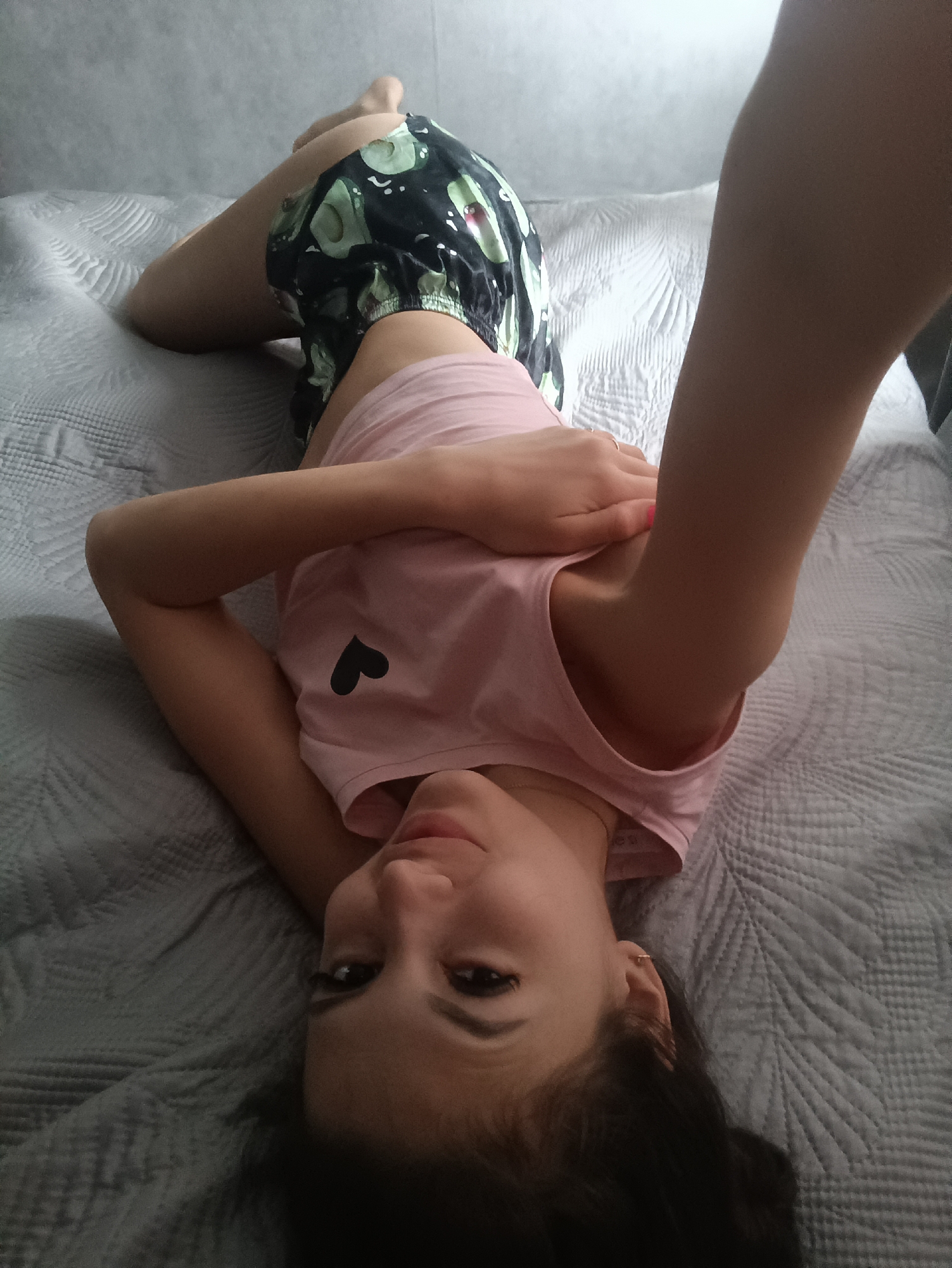 A young woman is lying on a bed. She is wearing a pink top with a black heart on it and black shorts with avocado patterns. The bed has a gray quilted blanket. She is looking at the camera and has one arm raised.