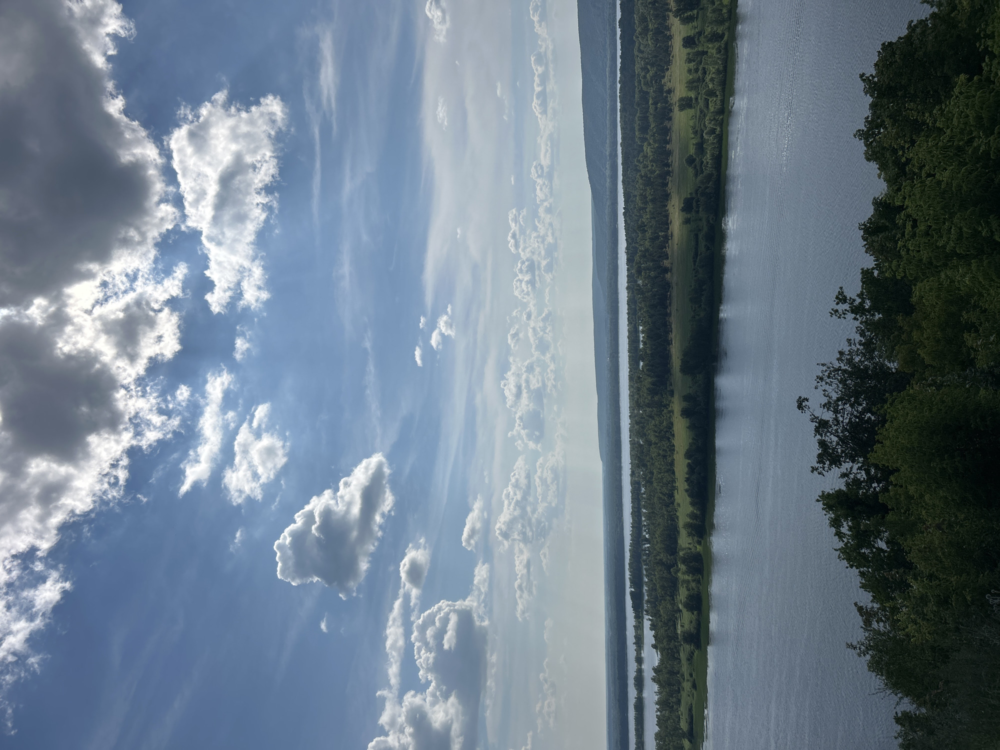 The image captures a wide, expansive river flowing through a lush green landscape under a dramatic sky filled with both bright white and dark gray clouds. The foreground is dominated by the dark green foliage of trees. The middle ground features the wide expanse of the river, with smaller islands or peninsulas covered in dense trees and green fields. In the distance, a hazy shoreline with a gentle slope can be seen, with the river appearing to widen as it stretches towards the horizon. The sky is a vibrant blue, with sunbeams filtering through breaks in the clouds, creating a sense of depth and a bright, atmospheric lighting. The scene is natural and serene, with no visible signs of human activity. The location is identified as the Samara region of Russia, near Novokuybyshevsk, likely a viewpoint overlooking a large river such as the Volga.