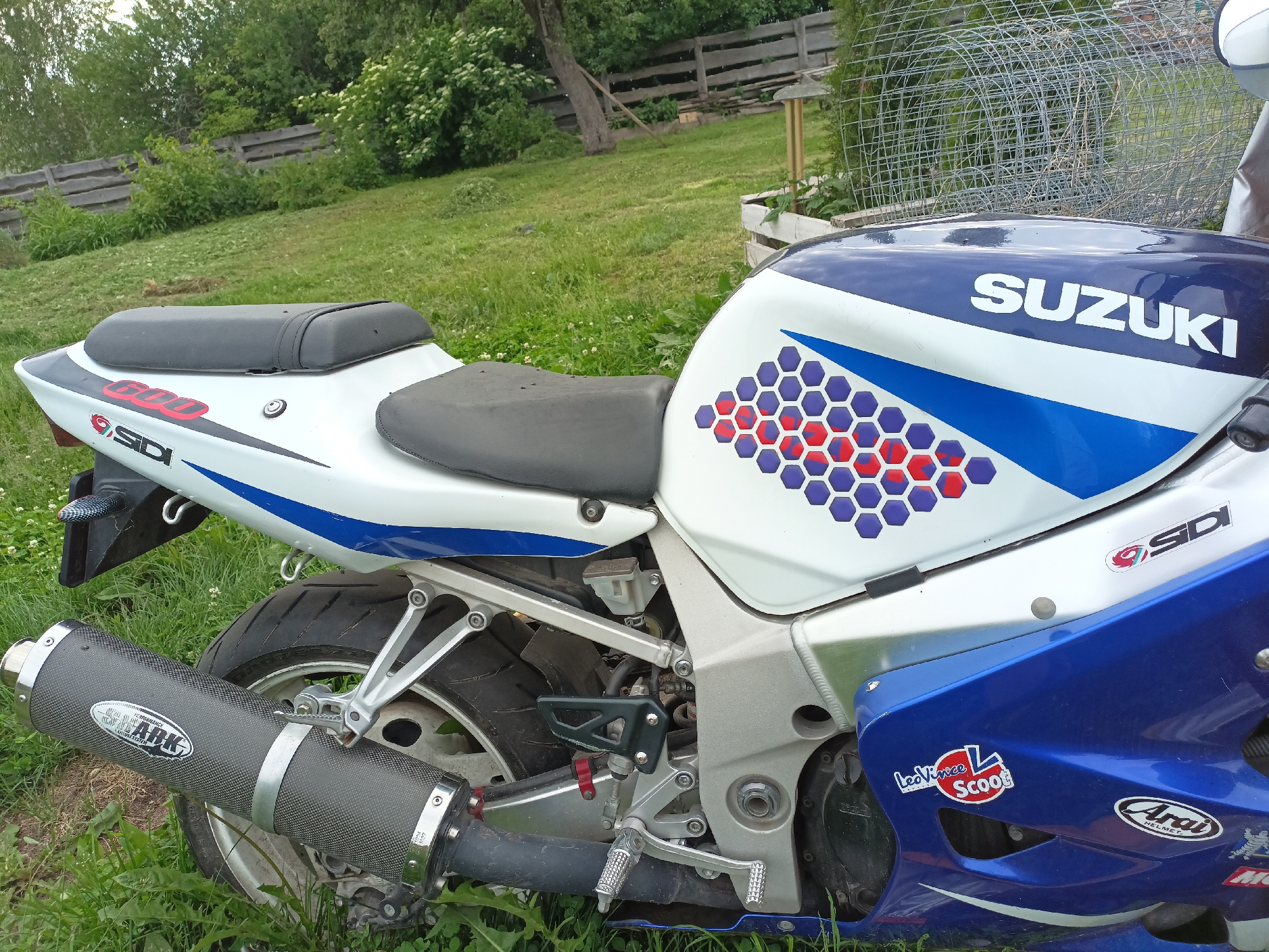 A blue and white Suzuki motorcycle is parked on a grassy area, possibly a yard or field, with a wooden fence and trees in the background. The motorcycle has "SUZUKI" written in white on the fuel tank, along with a hexagonal design in red and purple. It also features "GSX-R 600" branding on the tail. The motorcycle has a black seat and a carbon fiber exhaust pipe. There are various stickers on the motorcycle, including "SDi," "Leo Vince Scoot," and "Arai." The environment appears to be outdoors and rural.