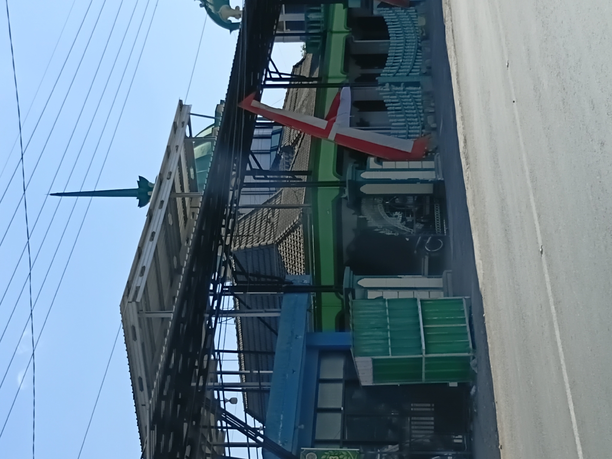 The image shows a building with a green and blue facade, partially covered by a corrugated metal roof supported by a dark metal frame. A tall, pointed green spire rises from behind the building. A red and white banner is draped diagonally across the front of the building. In front of the building, there is a light blue fence and a green metal enclosure. The scene is located on a street, with a road visible in the foreground. The sky is clear and blue, with power lines crossing the top of the image. The overall impression is of a mosque or religious building in an urban or semi-urban setting in Kasiyan Timur, Indonesia.