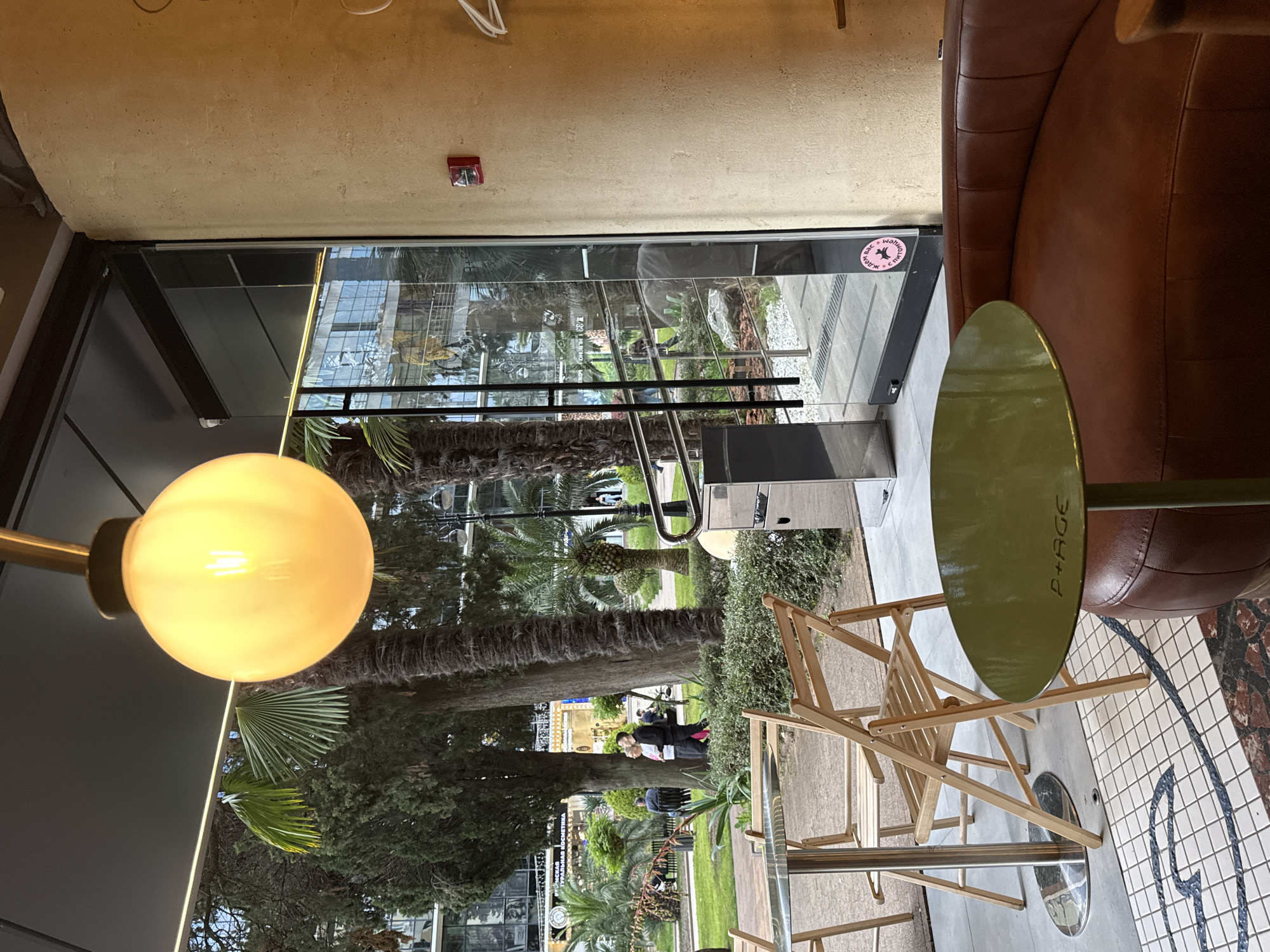 The image shows a partially outdoor scene with a view to a garden in what is possibly a restaurant setting. Through a glass door with a "Two Seasons Sochi" sticker, one can see an outdoor area with a garden, palm trees, and other greenery. Several individuals can be seen walking through the garden.
Inside, there is a round olive-green table with the "P+AGE" logo. A wooden folding chair is placed at the table, which sits near a brown leather sofa. The floor features a patterned mosaic. The interior ceiling is a light tan color. A modern lamp with a large, round, glowing bulb is visible. A fire alarm is attached to the ceiling.