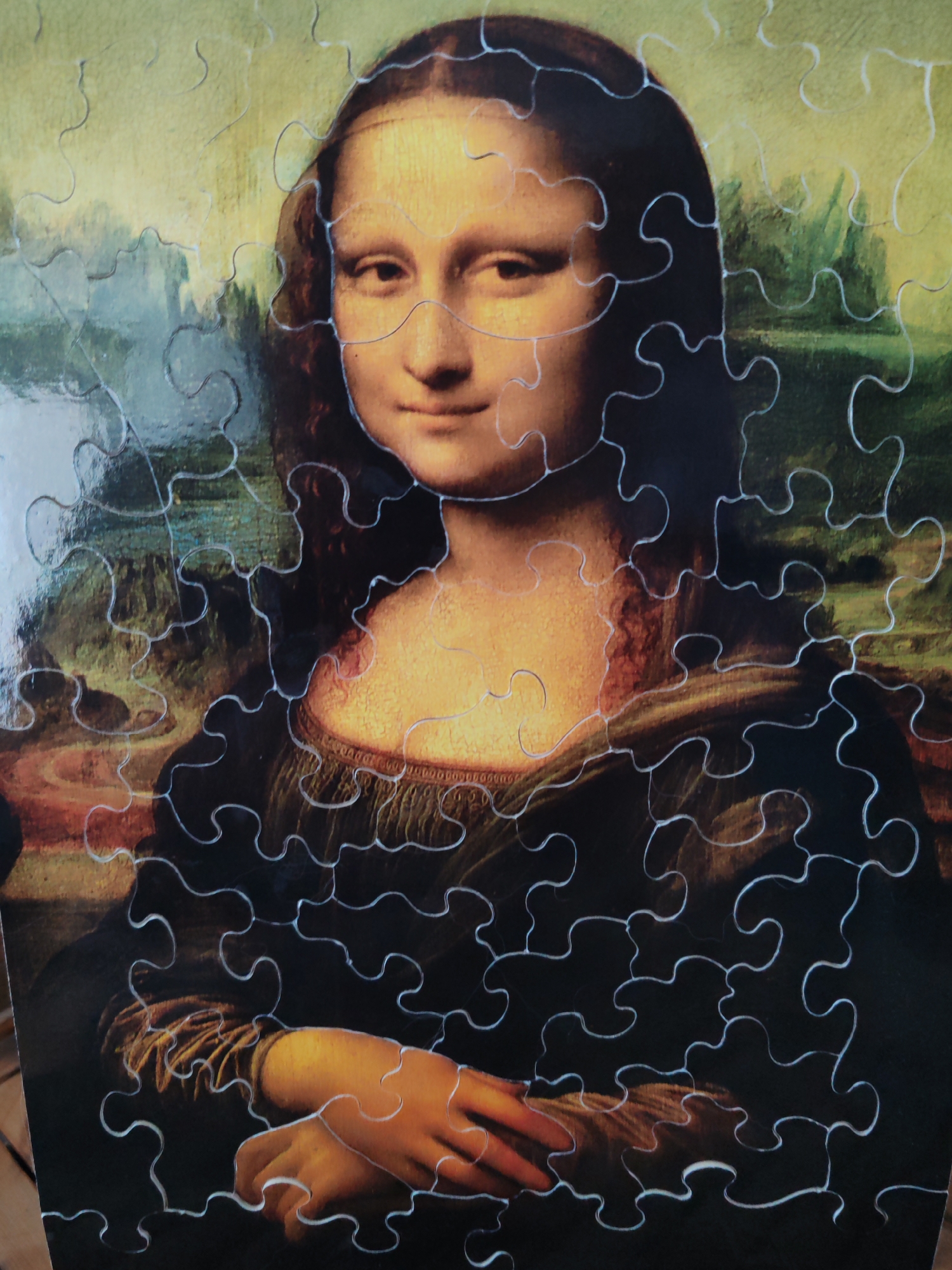 The image is a close-up, overhead shot of a jigsaw puzzle depicting Leonardo da Vinci's Mona Lisa. The puzzle pieces are clearly visible, forming the famous portrait of a woman with a subtle smile and enigmatic gaze. The background of the puzzle shows a hazy, dreamlike landscape. The puzzle is lying on a surface that appears to be a wooden table, with a slight sheen indicating a polished finish. There is no indication of any specific location in Dubai, United Arab Emirates.