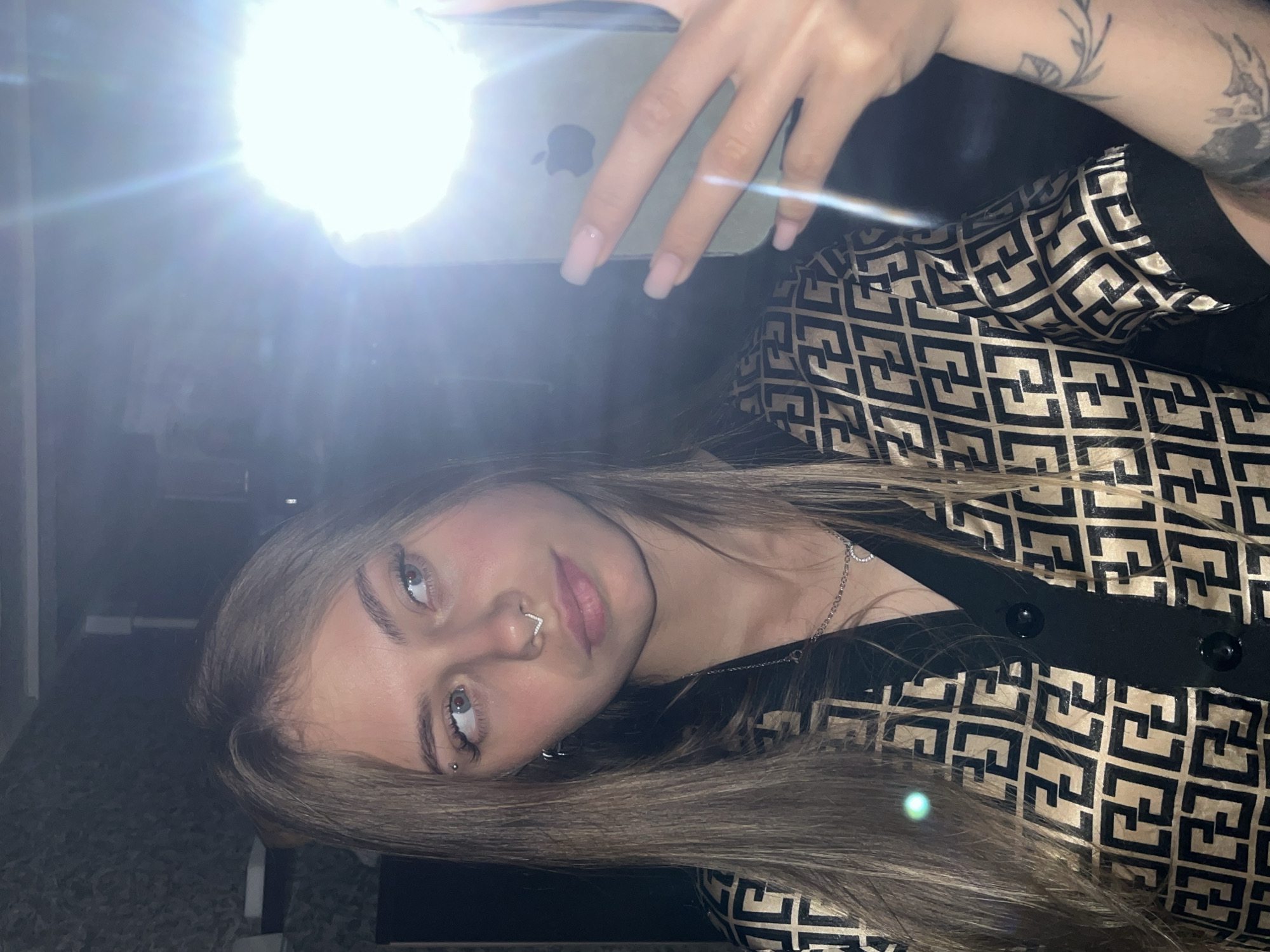 In Фоки, Russia, a young woman is captured in a mirror selfie, holding a light-colored iPhone with an illuminated flash. She has long, straight, light brown hair. Her eyes are light-colored, and she wears an eyebrow piercing on her left brow and a V-shaped septum piercing in her nose. She has light pink or nude colored, square-shaped fingernails. She is wearing a black and gold patterned shirt, possibly featuring a repeating "F" logo design, with a black collar and buttons. A silver-toned necklace with a small pendant is visible around her neck. A tattoo featuring botanical elements is visible on her left forearm. The background is dark, suggesting an indoor setting with minimal ambient light, largely illuminated by the phone's flash.