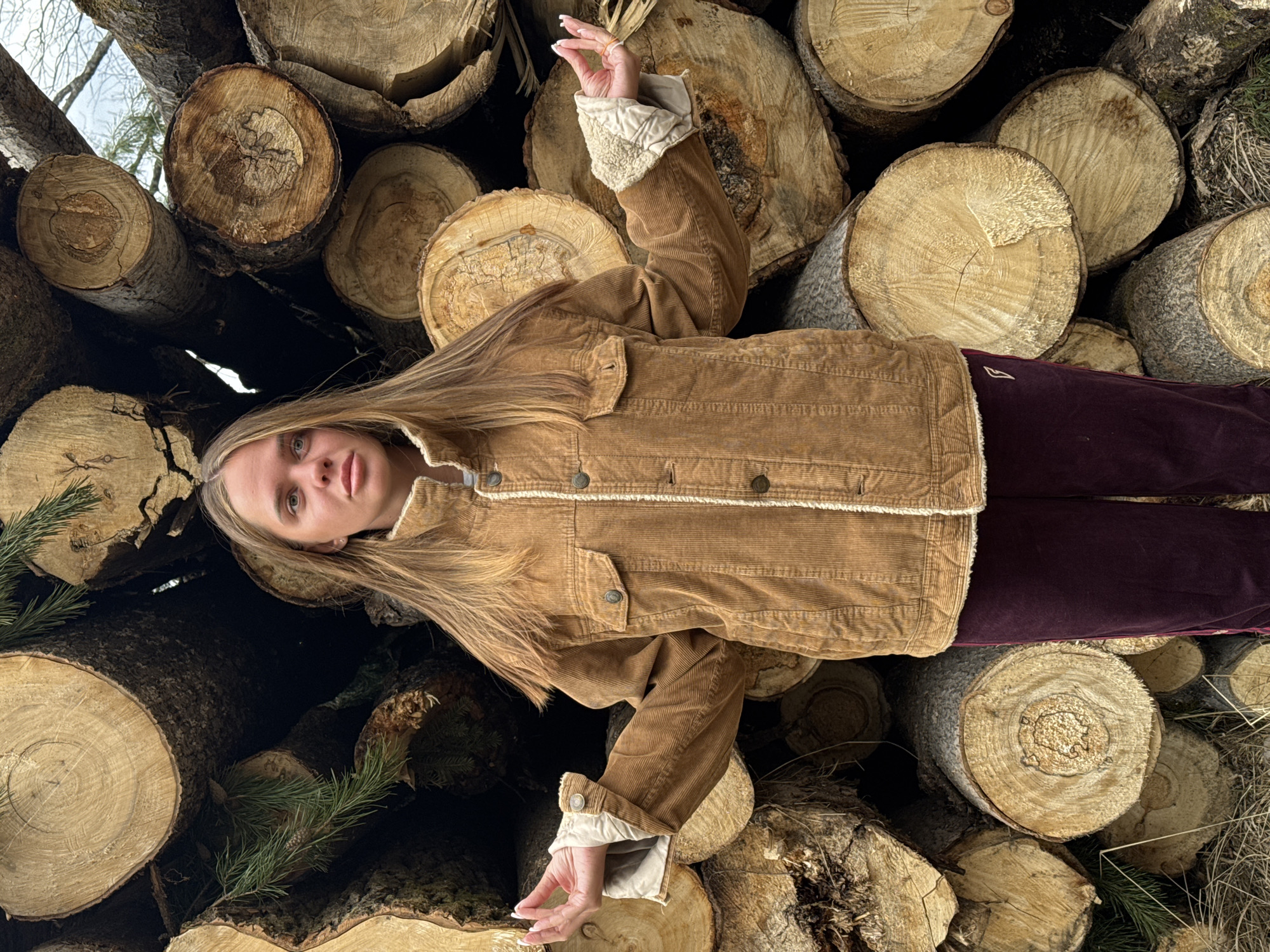 The photograph depicts a woman lying on a pile of logs. She is wearing a brown corduroy jacket with a white shearling lining and burgundy velvet pants. Her long blonde hair is spread across the logs. She looks directly at the camera. Her right arm is extended upward, hand slightly curved. The logs vary in size and color, with some showing tree rings. Sprigs of evergreen branches are interspersed among the logs. Based on the composition, location in the city of Grebnevo, Russia is not confirmed. No evidence suggests the presence of Grebnevo.