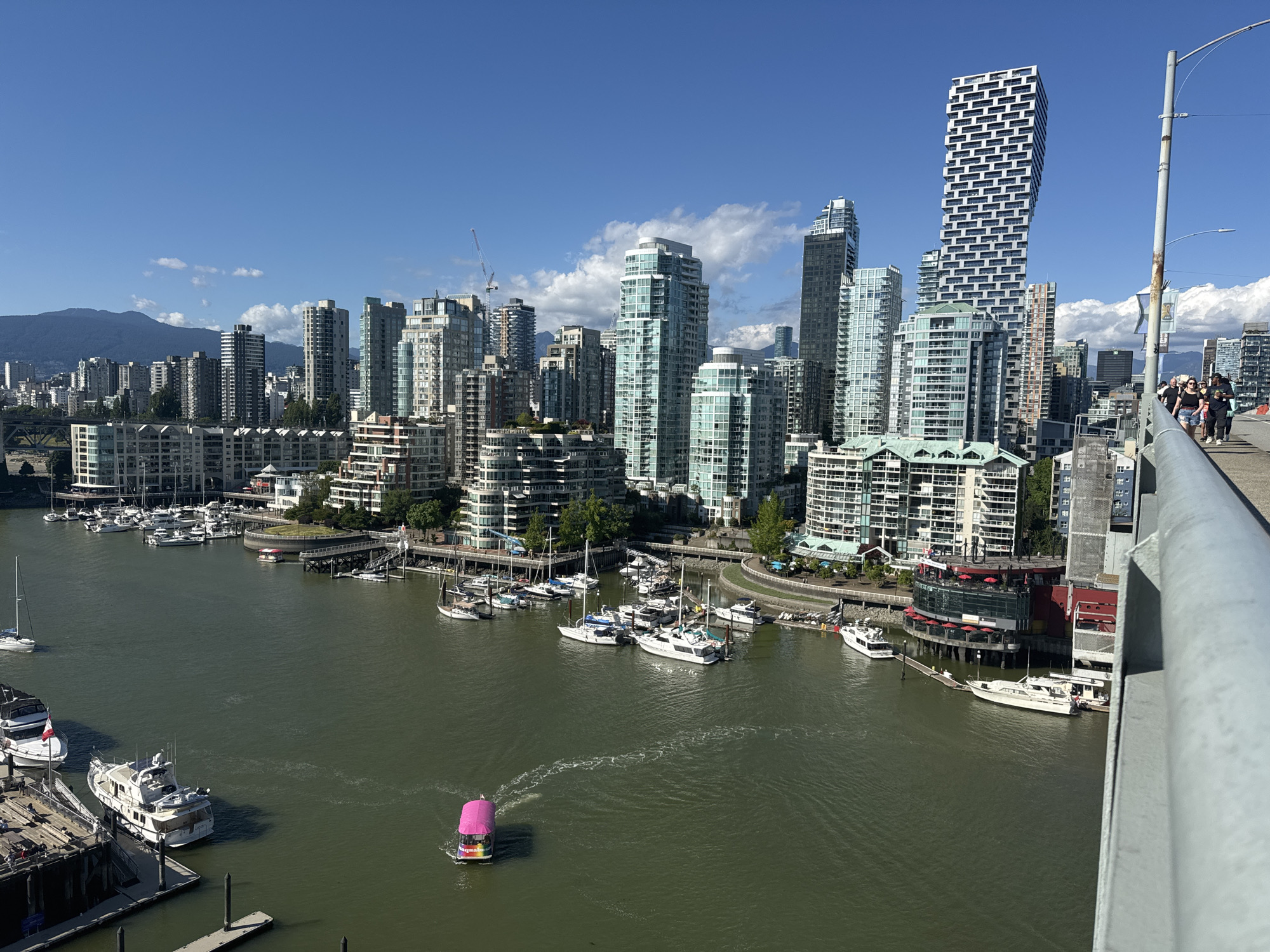 The image shows a wide view of the Vancouver waterfront, with the downtown skyline and Coal Harbour visible. In the foreground, a wide, green body of water is dotted with various boats, including sailboats, yachts, and a small pink vessel with a colorful exterior. Some of these boats are docked along wooden piers, while others are out on the water.
The middle ground features a marina filled with numerous sailboats and yachts. Beyond the marina, a strip of waterfront buildings with balconies and green spaces lines the shore. The Vancouver skyline dominates the background, with a cluster of modern skyscrapers, including the distinctive stacked-block building known as the "Living Shangri-La" and the twisting "Vancouver House." The buildings are a mix of glass and concrete, reflecting the bright blue sky with scattered white clouds. In the distance, a hazy mountain range is visible.
On the right side of the image, a pedestrian walkway is visible, with several people walking along it. A metal railing, likely part of a bridge or elevated walkway, runs along the edge of the frame. The overall scene is one of a bustling urban waterfront on a clear, sunny day.