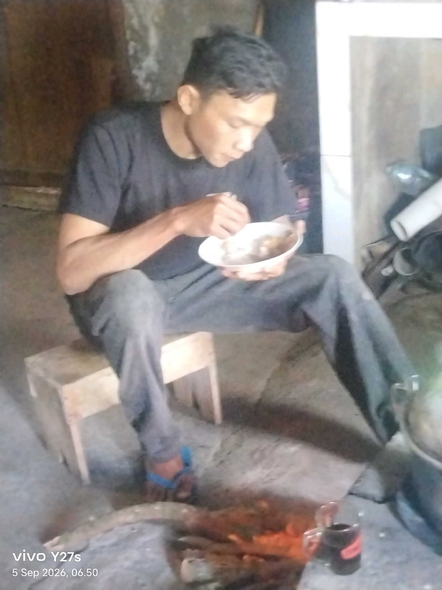 A man sits on a wooden stool, eating from a bowl. He is wearing a dark t-shirt and dark pants. In front of him, a fire burns on the ground, with a glass of dark liquid nearby. The scene appears to be indoors, with a rough, textured wall behind the man. The image is timestamped "5 Sep 2026, 06.50" and displays "vivo Y27s" in the lower left corner, suggesting it was taken in Tamansari, Indonesia.