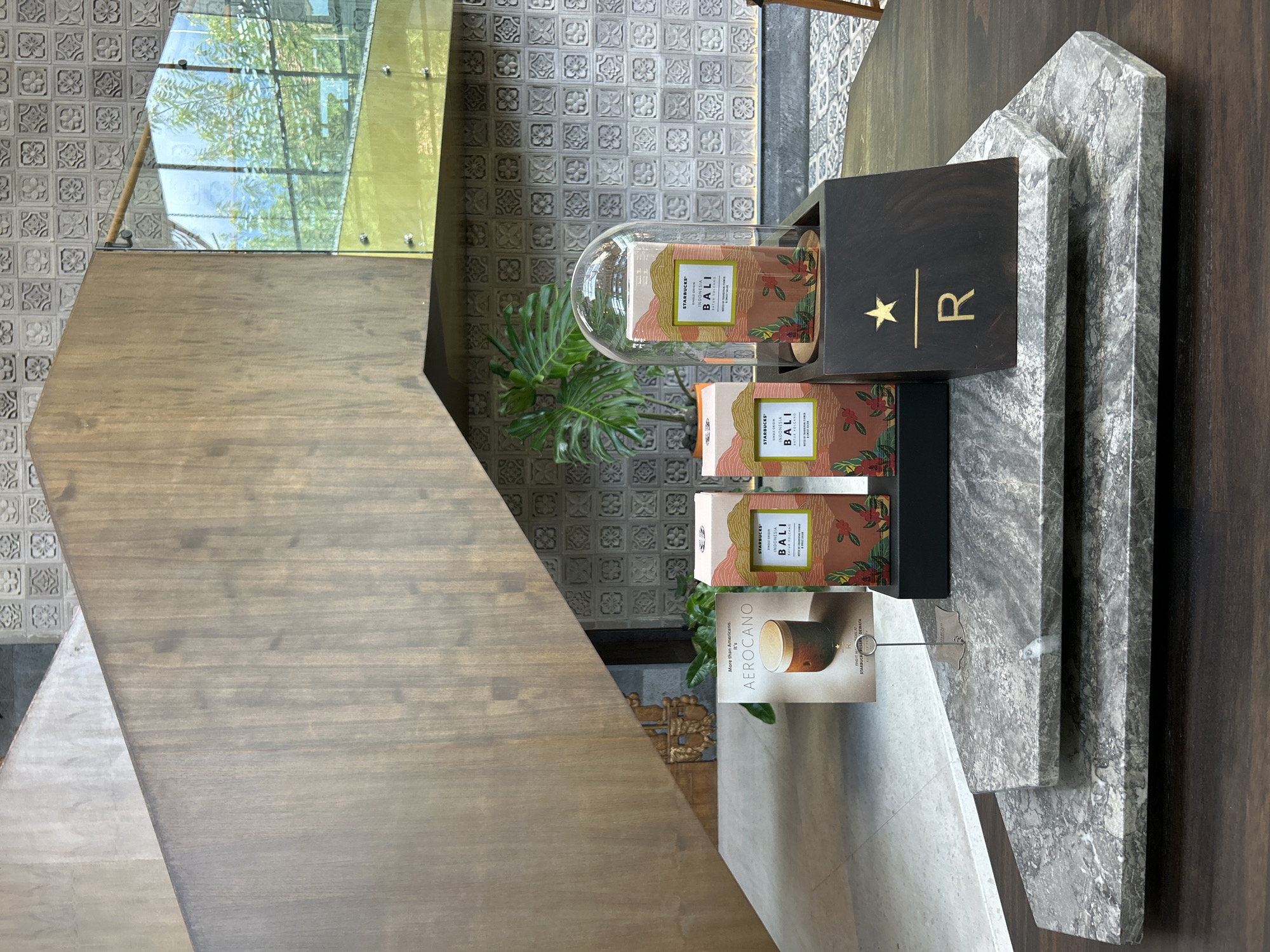 The image depicts a display of Starbucks Reserve coffee products, likely within a Starbucks Reserve store or a designated area within a regular Starbucks in Kuta, Indonesia. The arrangement is set against a backdrop of textured grey tiles and a modern wooden staircase with glass railings.
In the foreground, two hexagonal marble platforms are layered, with the upper one supporting a dark wooden display block. This block features the Starbucks Reserve logo: a golden star above a horizontal line and the letter "R". On top of the display block, a glass dome encloses a box of Starbucks Reserve "INDONESIA BALI" coffee. This box is predominantly coral pink and green, featuring floral and mountainous imagery and text identifying it as "SINGLE ORIGIN," "BALI," and "KARANG VOLCANO." To the left of this domed display, another identical box of "INDONESIA BALI" coffee stands on a black riser.
Further to the left, a brochure or book titled "AEROCANO" is propped up. It features an image of an Aerocano drink. A small metal stand holds this brochure. Behind these items, a large potted plant with broad green leaves is visible, adding a touch of nature to the scene.
The overall aesthetic is modern and sophisticated, with a focus on natural materials like wood and marble, and the presentation of premium coffee products. The Kuta, Indonesia location is suggested by the Starbucks Reserve offerings that might be specific to the region or country.
