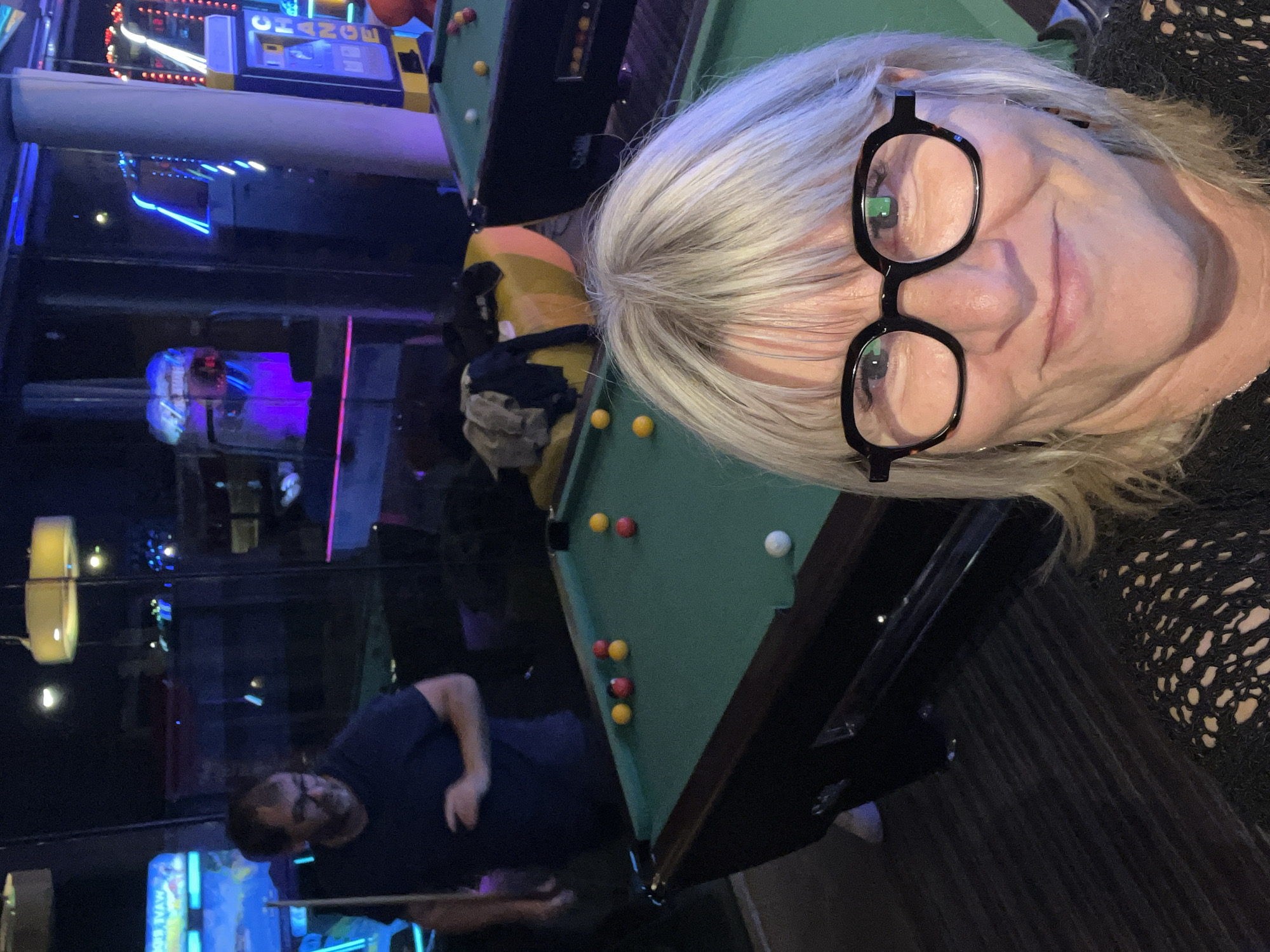 The image depicts a selfie of a woman with short, gray hair and black-framed glasses, taken indoors in what appears to be an arcade or amusement center. She is positioned near a pool table with several billiard balls scattered across the green felt. A man is visible in the background, holding a pool cue and seemingly preparing to take a shot. Various arcade games and illuminated displays are also discernible in the background, adding to the ambiance. Based on this information, the location is Montrond-les-Bains, France.