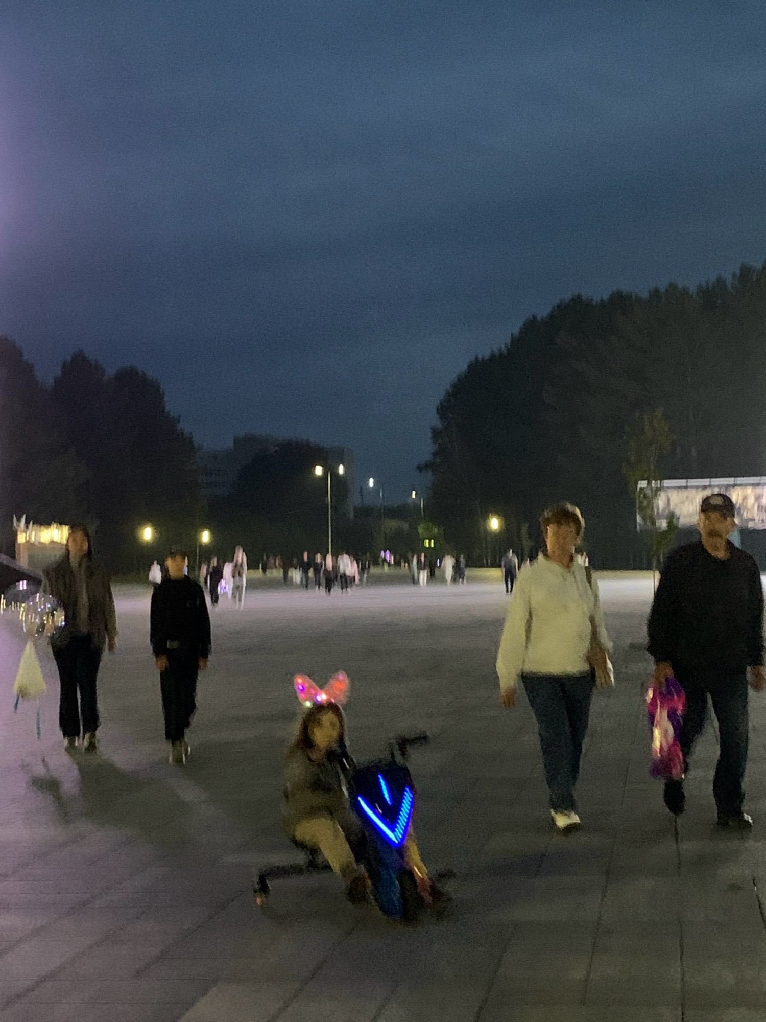 In Prokopyevsk, Russia, an evening scene unfolds on a paved outdoor area with scattered trees in the background. Several individuals are visible. In the foreground, a child wearing illuminated bunny ears is seated on a blue and yellow electric scooter. To the right, an adult male and female are walking. The male is wearing a dark jacket and a baseball cap, carrying a bag with colorful tissue paper. The female is wearing a white jacket and blue jeans. Further in the background, other people are walking, and streetlights illuminate the area. The sky is dark, indicating it is nighttime.