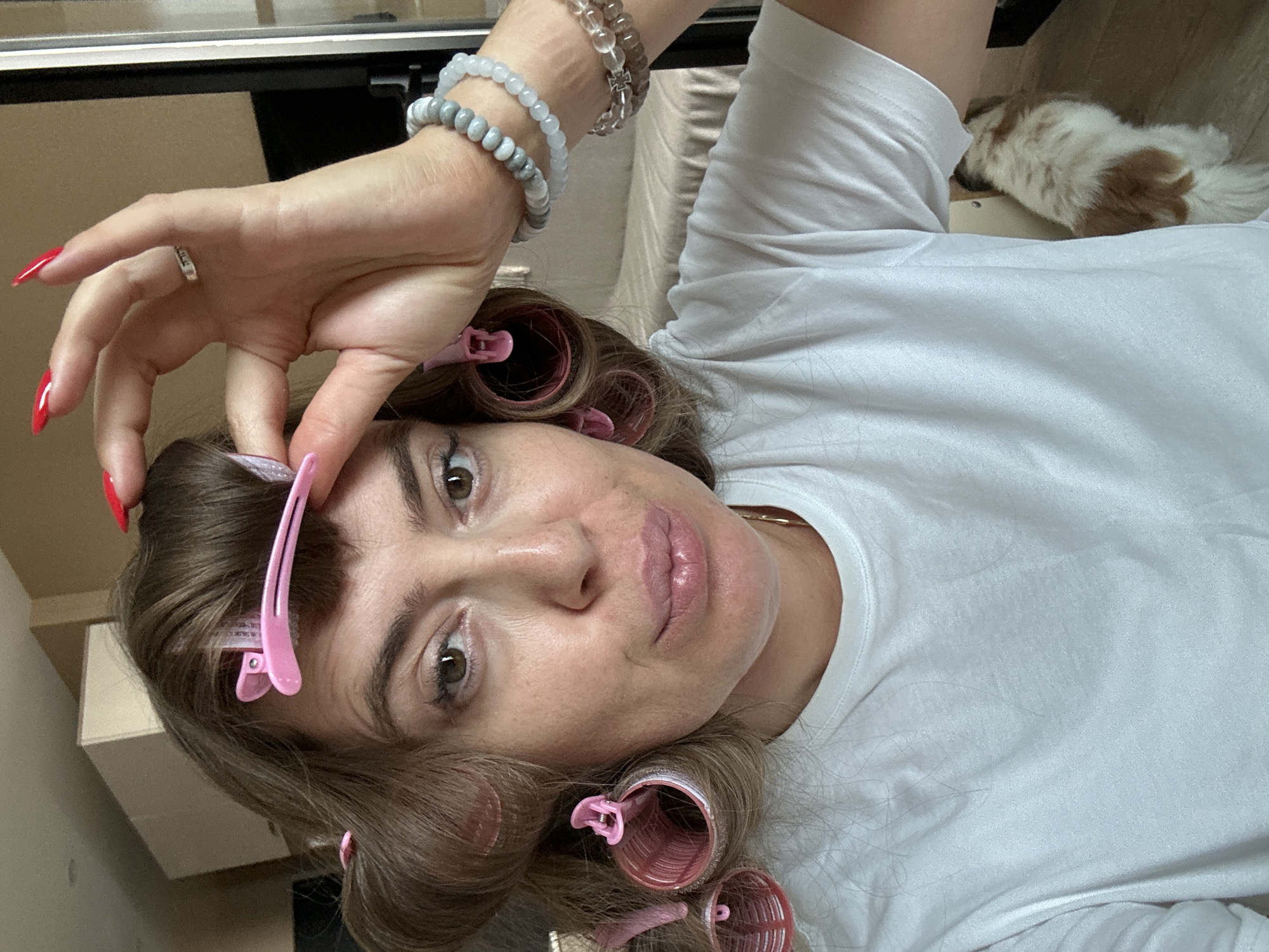 A woman is shown indoors, close-up to the camera, with hair rollers in her hair and bright red nail polish. She is wearing a white t-shirt. Her left hand is raised to her head, adjusting a roller. Her right arm is down by her side, showing a stack of bracelets on her wrist. Behind her, a dog with brown and white fur is visible peeking out from under a piece of furniture. The background suggests an indoor living space with a beige couch and wooden flooring. There are no discernible features indicating the city of Batumi, Georgia, in the image.