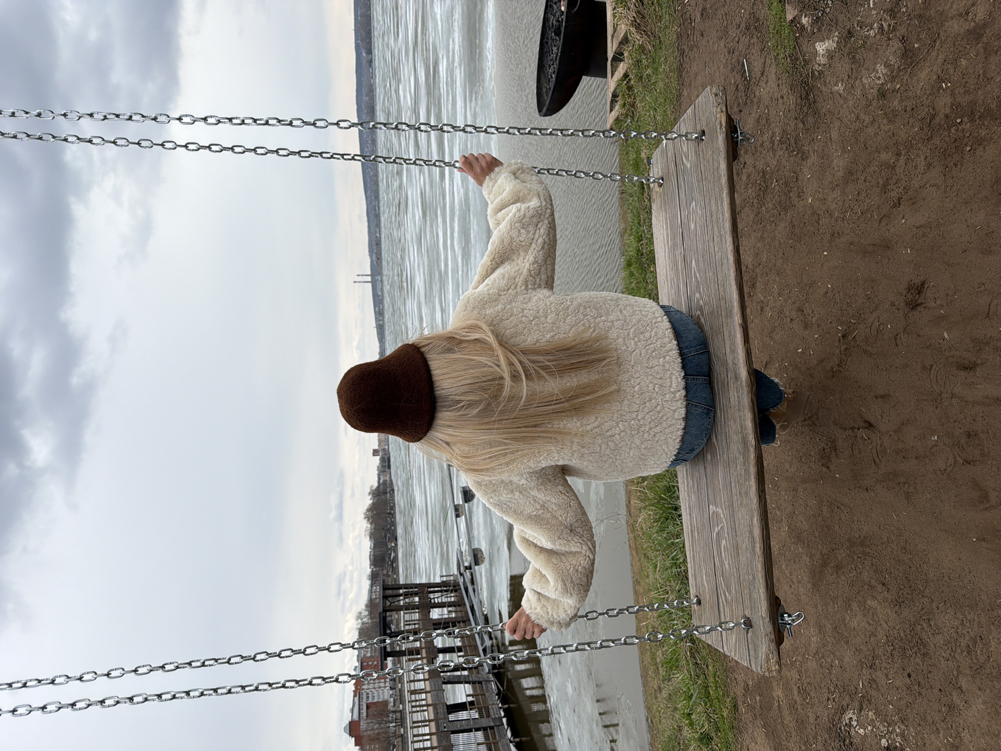 An overhead, slightly angled shot shows a person seated on a wooden swing, looking out over a wide body of water in Votkinsk, Russia. The person, seen from behind, has long blonde hair, wears a dark brown hat, a cream-colored fuzzy jacket, and blue jeans. Their hands grasp the metal chains of the swing.
The swing is positioned on a bank with brown dirt and patches of green grass. To the right of the swing, a black, round, bowl-like object (possibly a fire pit) sits on the grass. To the left, wooden piers or structures extend into the water near the bank. The body of water, likely the Votkinsk Reservoir, appears calm but somewhat textured, reflecting the overcast sky. On the distant horizon across the water, land is visible with faint outlines of trees, buildings, and two thin, tall industrial structures or chimneys.
The sky is uniformly grey and cloudy, suggesting an overcast day. The overall scene conveys a sense of peaceful contemplation or relaxation as the person sits and gazes at the expansive natural landscape. There is no visible text in the image.