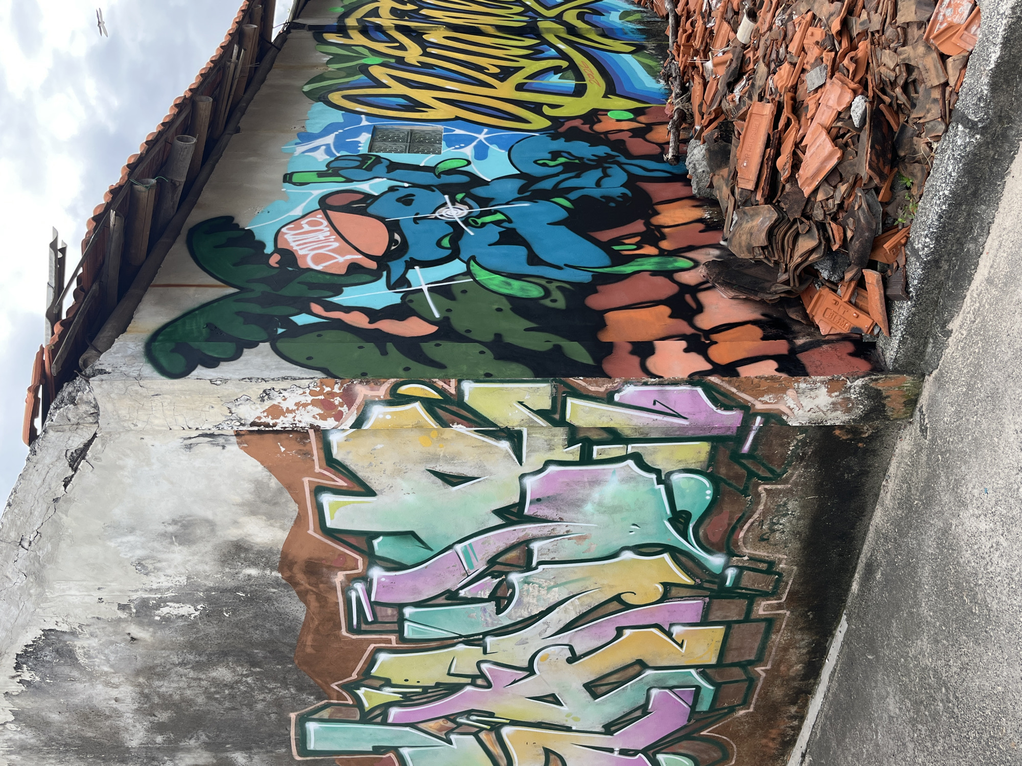 The image depicts a crumbling wall in Kuta, Indonesia, adorned with vibrant graffiti. The wall itself appears weathered and aged, with peeling paint and visible cracks. On the left side, graffiti featuring bubble letters in shades of purple, pink, yellow, and green covers a significant portion of the wall. To the right, a colorful mural dominates another section of the wall. This mural includes a blue cartoonish character with large ears and an orange hat, holding a green object and pointing a finger upwards. Behind the character, large green palm-like leaves are depicted. Further to the right, more graffiti with yellow and blue lettering is visible. The roof of the structure is made of clay tiles, some of which are in disrepair. A pile of broken clay roof tiles lies on the ground in front of the wall. The ground is paved with asphalt. The sky is overcast with a few distant white objects visible. The scene suggests an urban environment that is either undergoing demolition or has been neglected, with street art being a prominent feature.