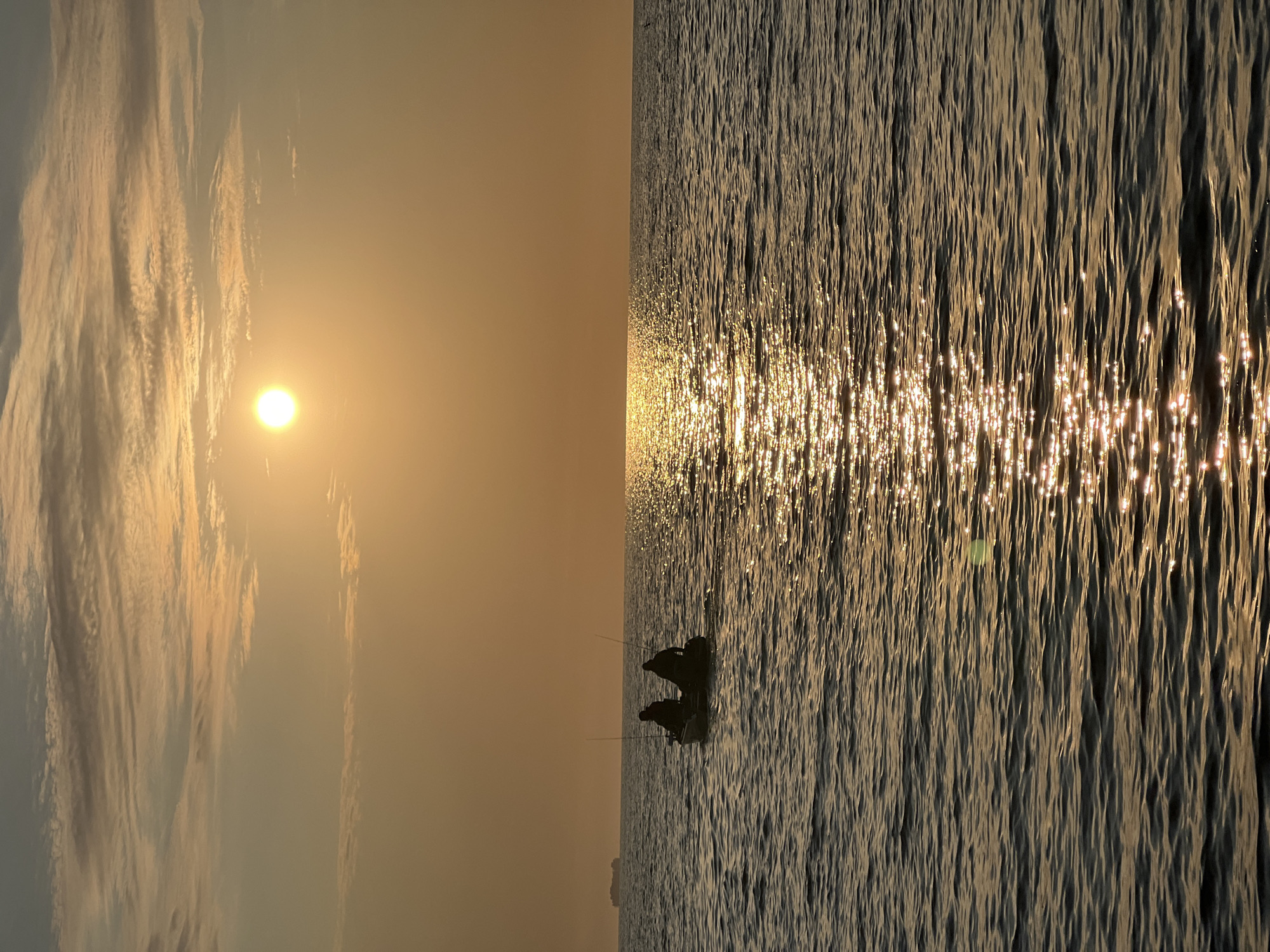 This is a photograph taken at sea, likely during sunset or sunrise, given the warm, golden hues of the sky and the reflection of the sun on the water. In the mid-ground, a small inflatable boat is visible, carrying two individuals who appear to be fishing. They are silhouetted against the bright sky. In the far distance, a large ship is faintly discernible on the horizon. The water is slightly choppy, with ripples catching the light. The sky is partly cloudy, with elongated cloud formations. The scene suggests a peaceful and serene moment at sea. The reference to Borovoye, Russia, appears to be a misunderstanding, as Borovoye is a resort town in Kazakhstan, not Russia, and the image depicts a marine environment, not the landscapes typically associated with Borovoye.