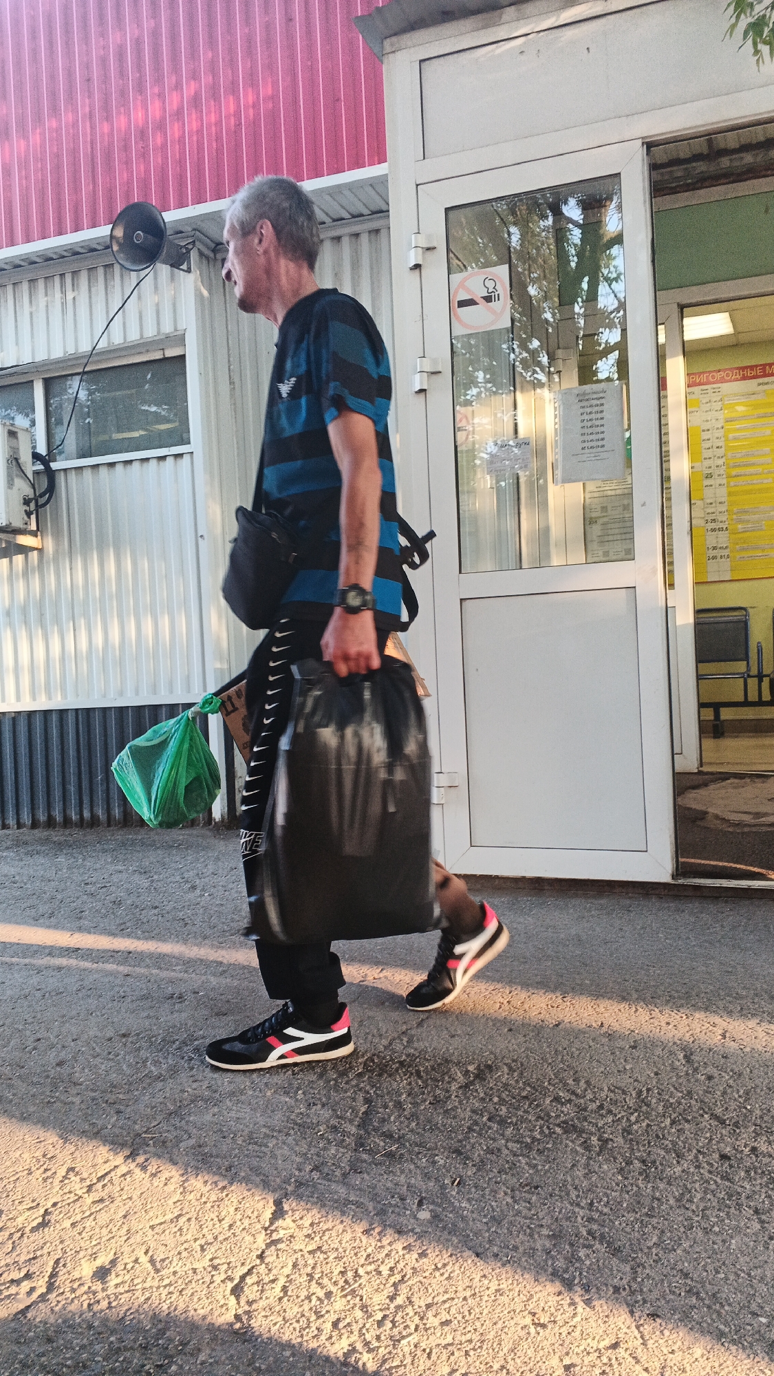 A man is walking outdoors in the city of Izhevsk, Russia, with a black shoulder bag, a green plastic bag, and a large black bag. He is wearing a blue and black striped t-shirt, black pants with white stripes, and black and white sneakers with pink accents. The man is walking on a paved surface in front of a building with a glass door and red corrugated metal siding. There is a "no smoking" sign on the glass door. The sun is casting long shadows on the ground.