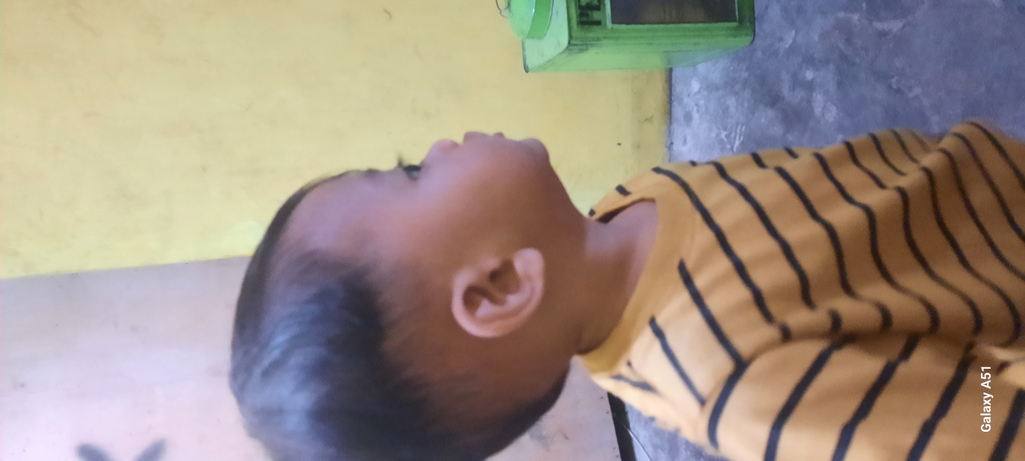 The image shows a close-up profile view of a young child, likely a toddler, facing right. The child has dark hair, a rounded face, and is wearing a yellow t-shirt with thin dark stripes. The background consists of a textured wall that appears to be painted yellow in some areas and a lighter, possibly pinkish or white color in others. To the right of the child, a green rectangular object with a lid is partially visible. A watermark in the bottom left corner of the image reads "Galaxy A51". The location is identified as Selopuro, Indonesia. The overall scene appears to be indoors, possibly in a residential setting, based on the wall texture and the presence of the green object which may be a storage container or bin. The lighting is somewhat dim, suggesting it could be evening or an indoor environment with limited natural light. There are no other people or specific actions depicted in the image besides the child's static pose.