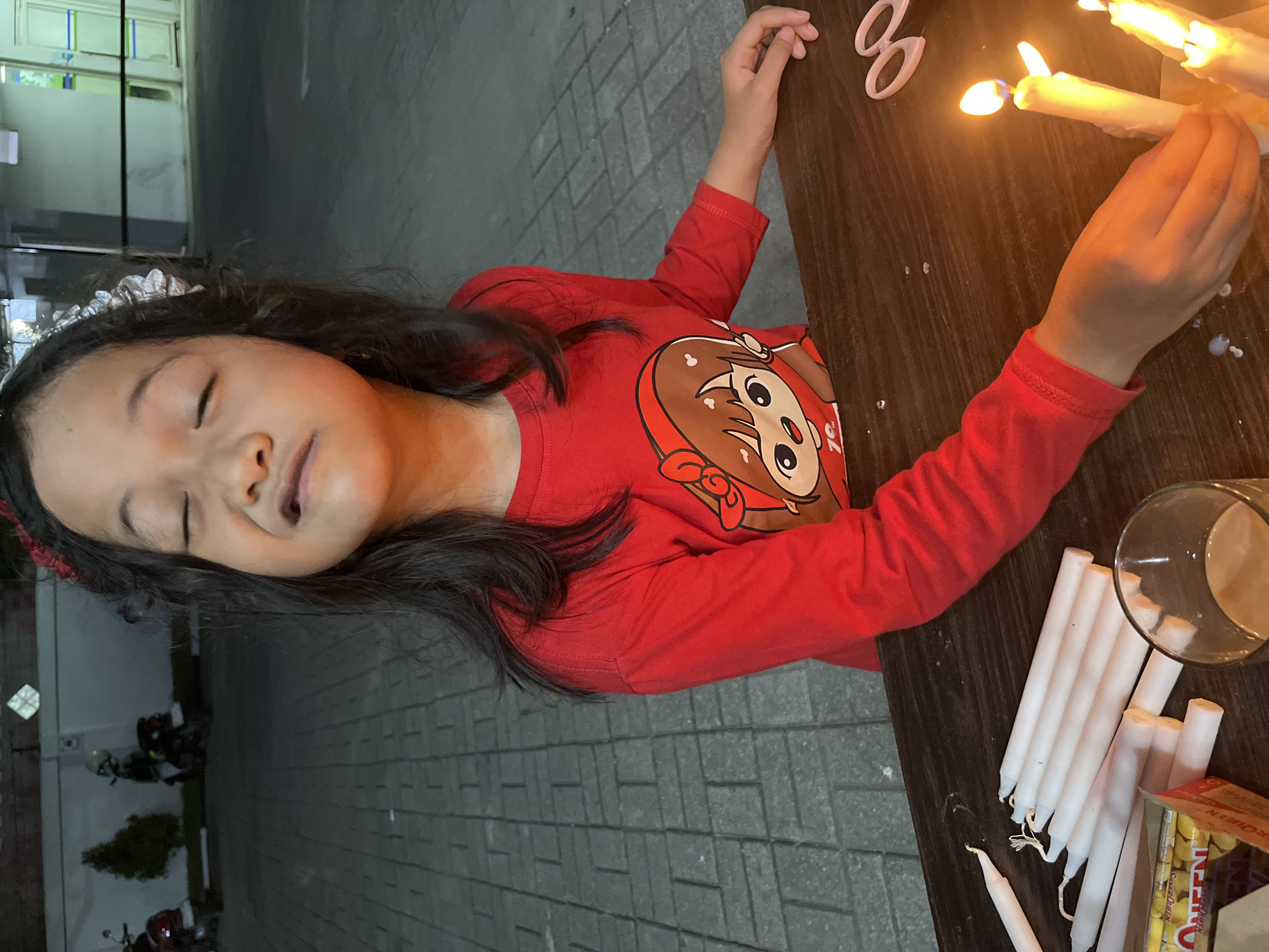 A young girl wearing a red long-sleeved shirt with a cartoon character print on it is seated at a wooden table, her eyes closed and face scrunched in an expressive grimace. She is adorned with a decorative red hair accessory. Before her, a collection of white candles are laid out on the table, some of which are lit and casting a warm glow. A glass containing a light-colored beverage sits nearby, along with a box of candy. In the background, the scene transitions to an outdoor environment with a paved walkway, a wall, and a few parked motorcycles. The overall impression is a candid moment captured during an activity that likely involves candles, possibly a celebration or a religious observance, set in a semi-urban environment in Kel. Pasangkayu, Indonesia.