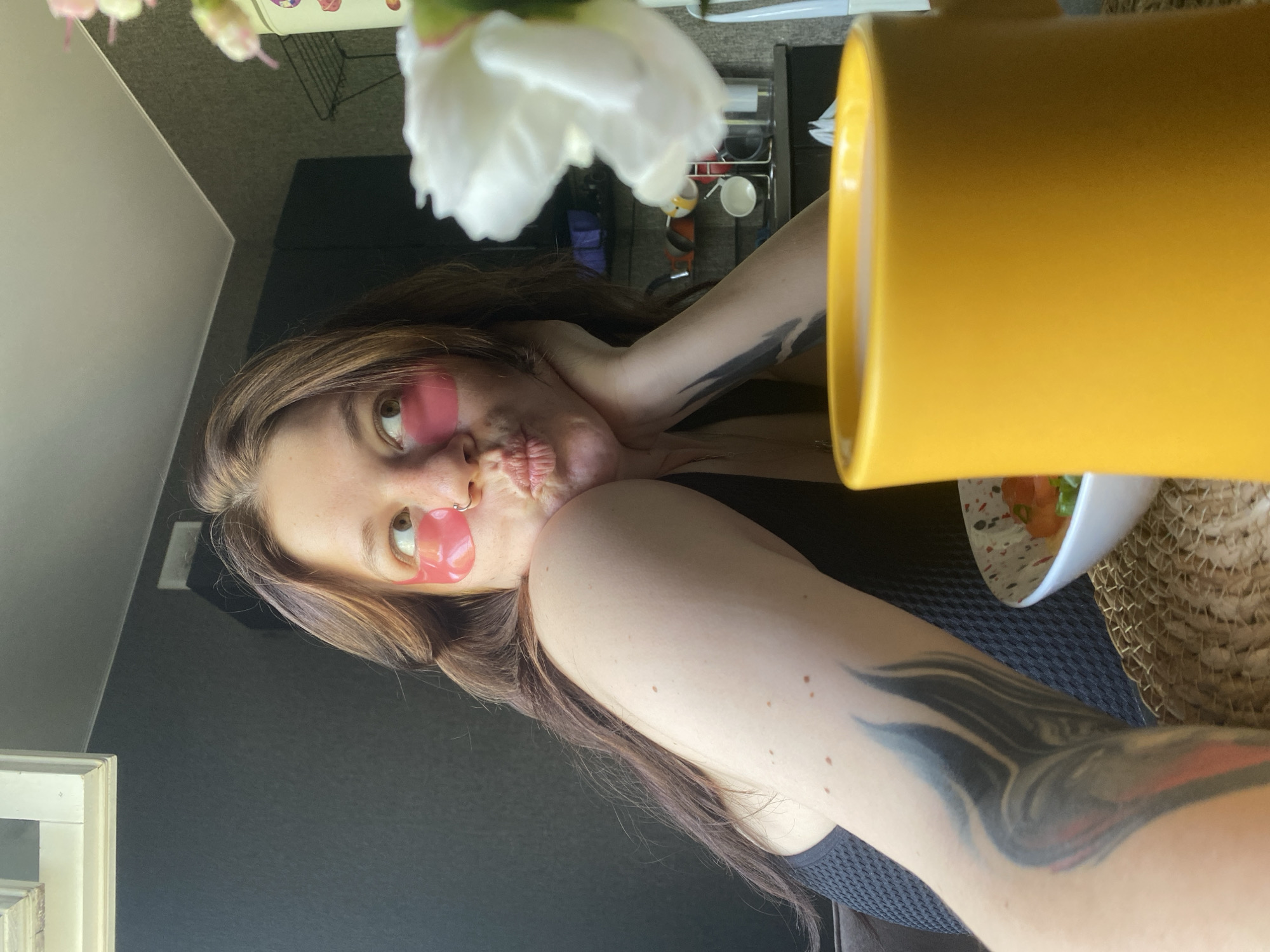 A young woman with brown hair is sitting at a table with a yellow mug and a bowl of food. She is wearing pink under-eye patches and a black top. There is a tattoo on her left arm. The background appears to be a kitchen or dining area. The image was taken in Chaikovsky, Russia.