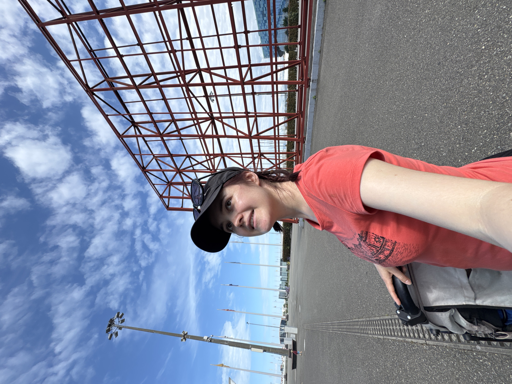 A woman is standing on a gray paved surface in the city of Sirius, Russia. She is wearing a coral-colored t-shirt and a black baseball cap, with sunglasses perched on top of the brim. The woman is smiling and looking off-camera to the left, possibly holding a stroller. Behind her, a large red metal grid structure rises against a blue sky filled with scattered white clouds. In the distance, a row of flag poles stretches along the horizon, interspersed with buildings. There are tall lighting poles.