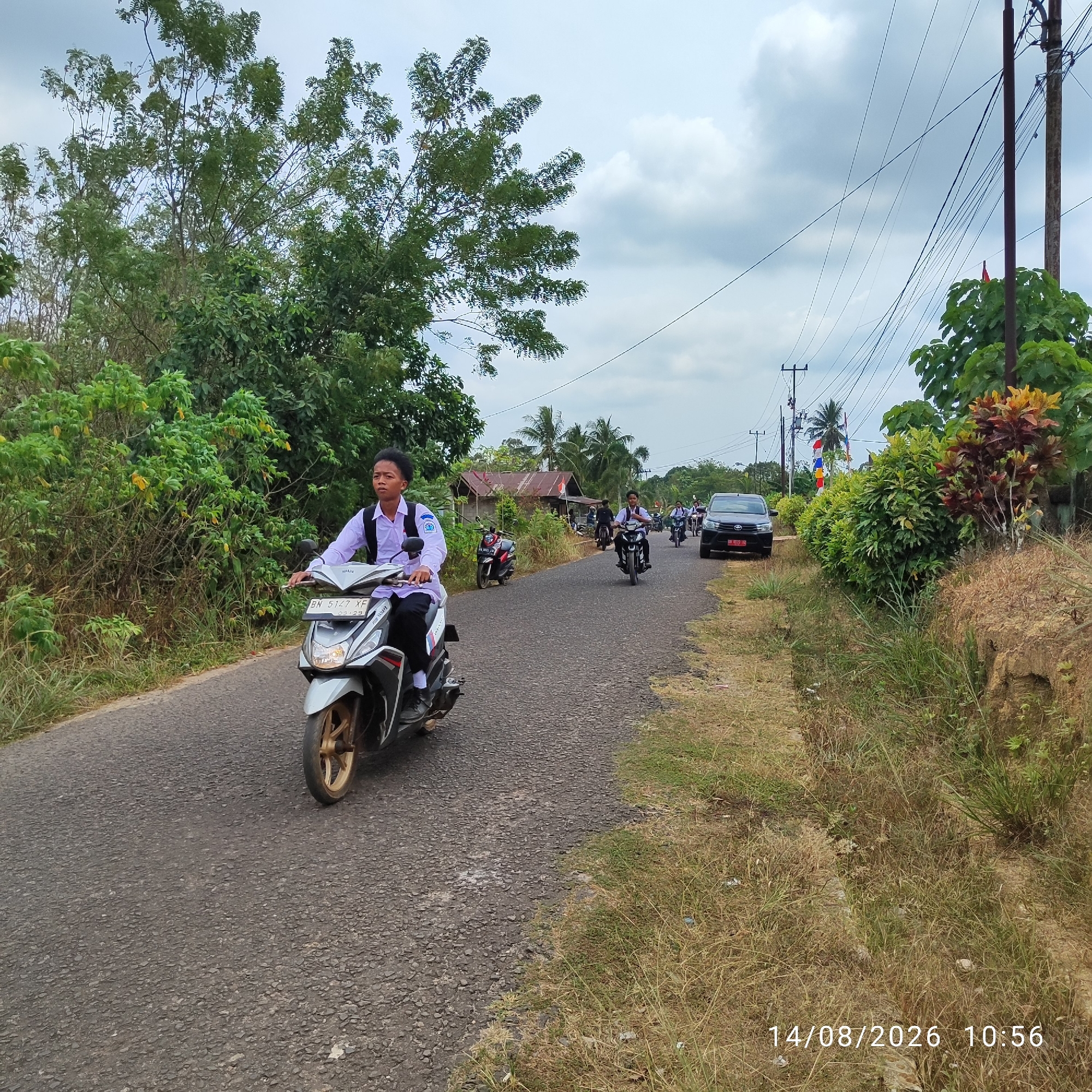 A group of students in uniform are riding motorcycles down a paved road in a rural area of Indonesia. The lead motorcycle is a silver scooter with a young male rider wearing a white shirt and dark pants, carrying a black backpack. He is looking forward as he rides. Behind him, other students on motorcycles are also visible, along with a dark colored SUV. The road is lined with lush green trees and vegetation on both sides. In the background, a small building with a rusty roof can be seen. Utility poles with numerous wires stretch across the sky. The sky is overcast with a few patches of blue. The date stamp "14/08/2026 10:56" is visible in the bottom right corner of the image.