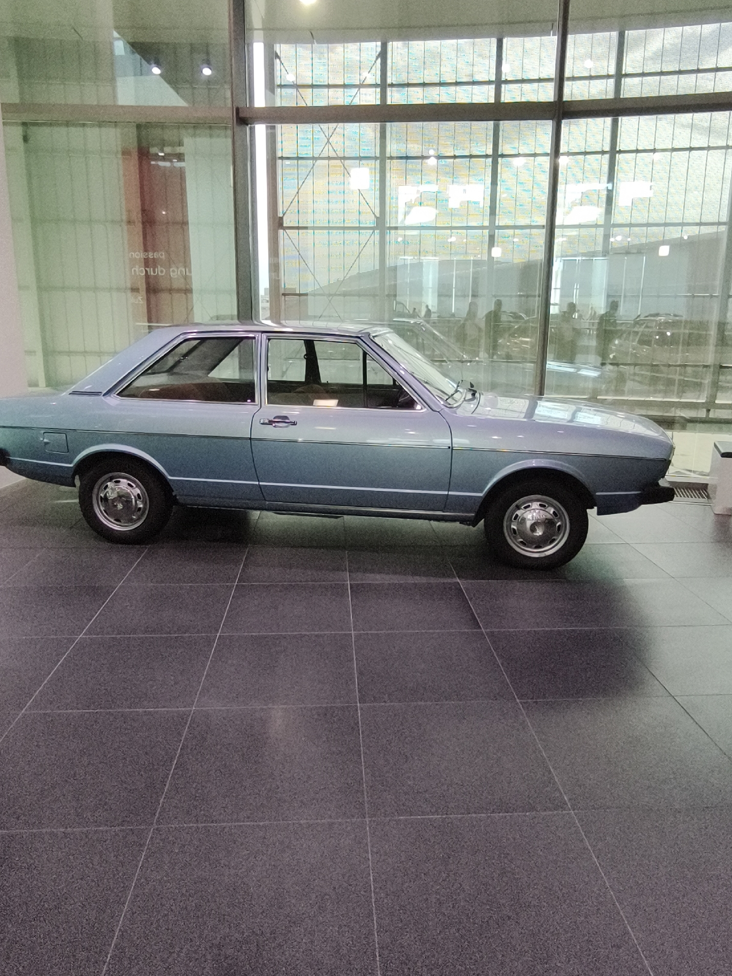The image captures the side profile of a light blue, two-door sedan displayed indoors on a dark tiled floor. The car appears to be an older model, possibly from the 1970s or 1980s, with a classic design. It is positioned in what seems to be a museum or exhibition space, suggested by the clean, well-lit environment and the large glass windows behind the vehicle. Through the windows, a multi-paned structure and reflections of other vehicles and figures can be observed. The car itself is the primary subject, showcasing its sleek lines, chrome wheels, and tinted windows. The environment is sterile and organized, typical of a museum setting dedicated to automotive history or design. It is likely located in Ingolstadt, Germany, given the city's strong association with the Audi brand and its prominent museum.