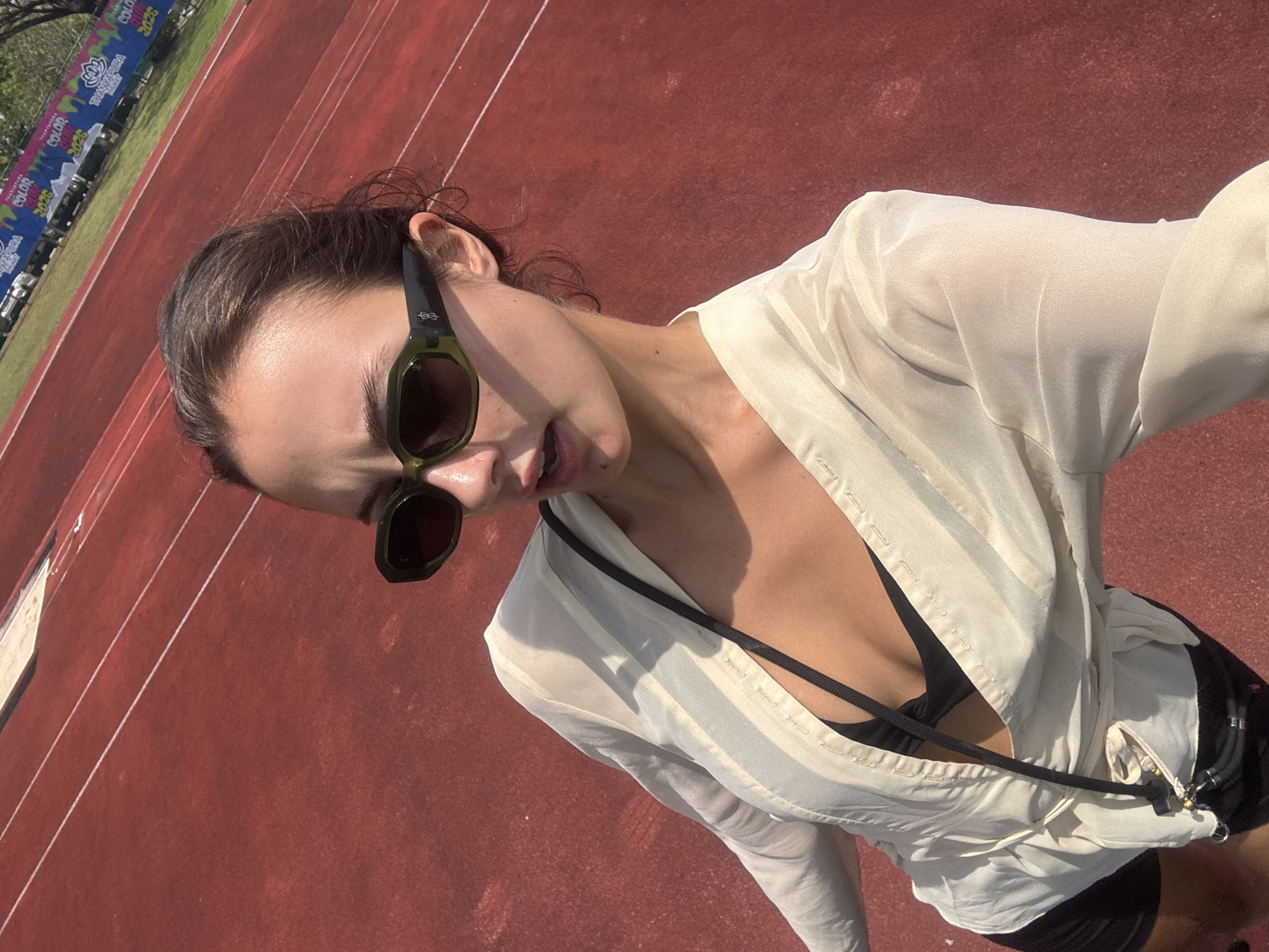 A woman wearing sunglasses and a light-colored blouse is standing on a red running track. The track is marked with white lines. The background includes banners with the words "COLOR THALANG MANORAMA 2025". The photo was taken outside in daylight. The location is Thep Krasatti, Thailand.
