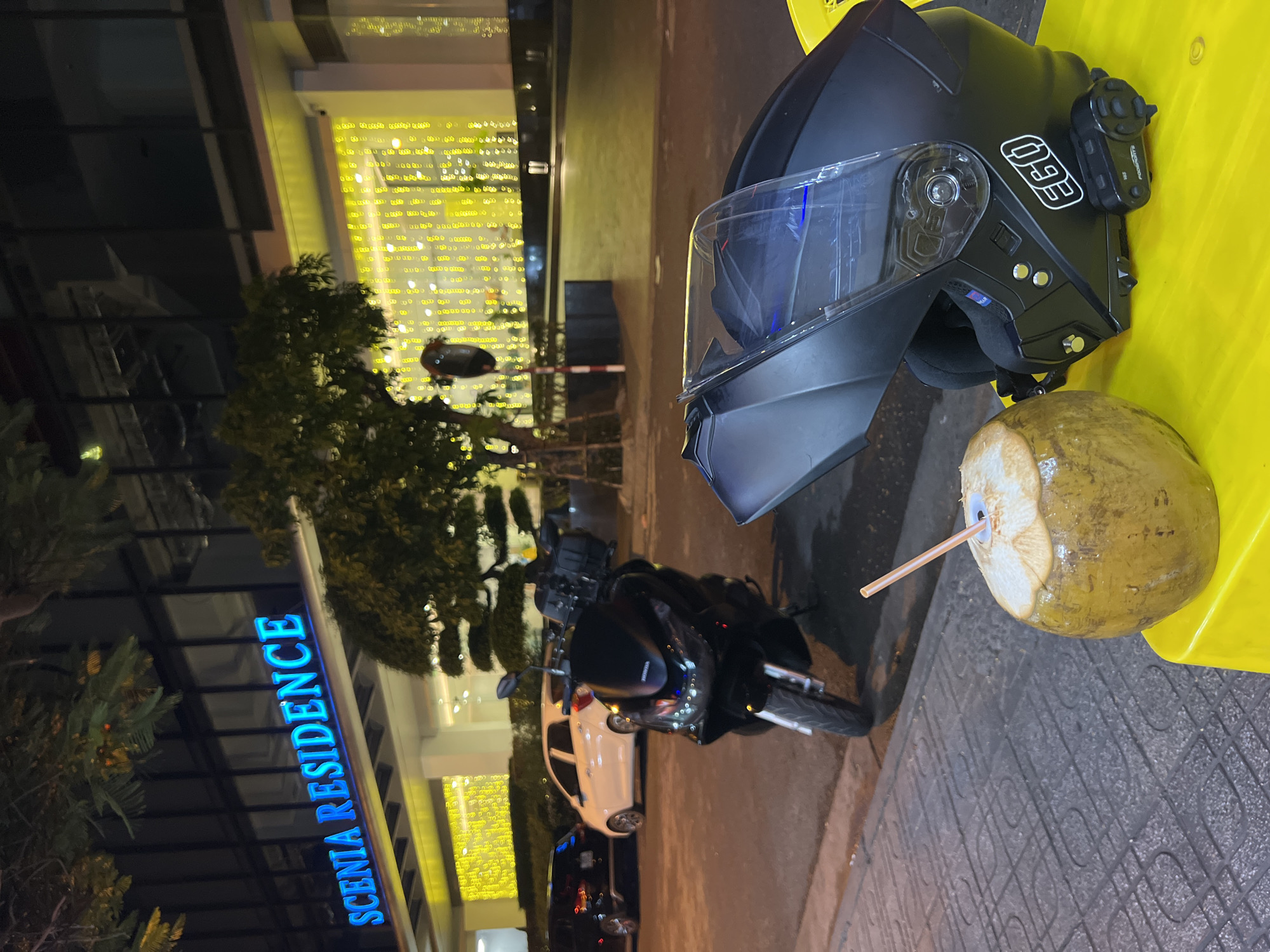 The image depicts a motorcycle helmet, a coconut with a straw, and a motorcycle parked outside a building identified as "SCENIA RESIDENCE". The scene appears to be at night, given the lighting conditions. The helmet is black with a clear visor and the logo "EGO" is visible. The coconut has a straw inserted, suggesting it's ready to drink. The building is brightly lit and the motorcycle is parked nearby. The location of the scene is possibly in the city of Bac Natural Heritage Area Trang, Vietnam.