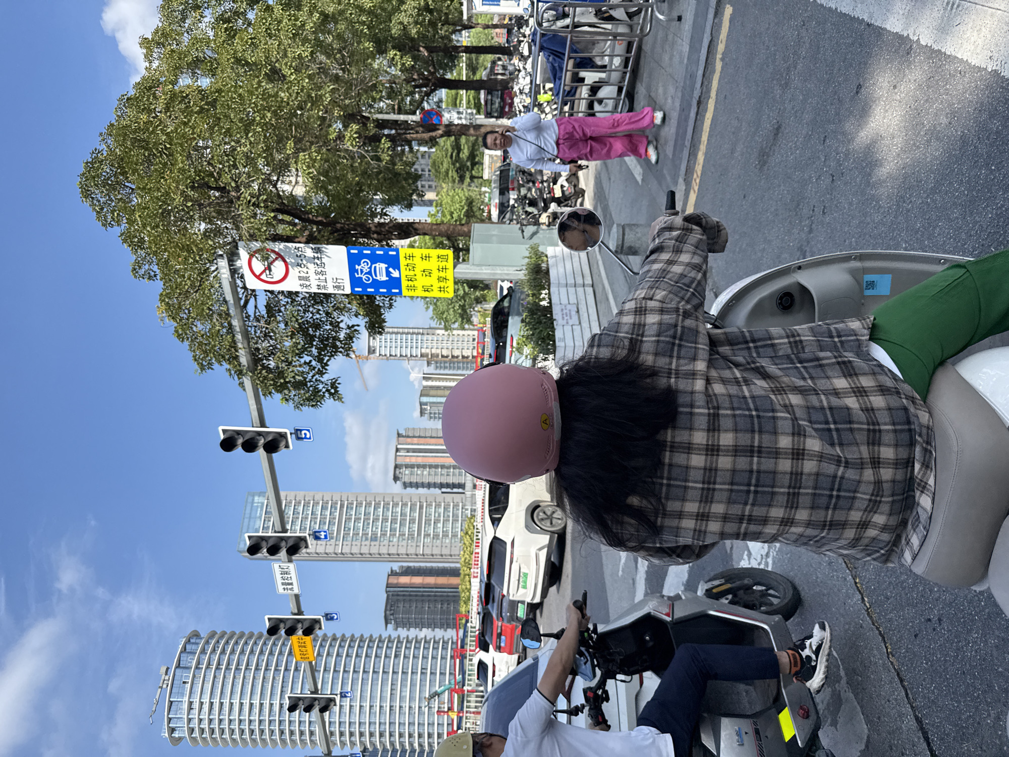 The image depicts a street scene in Tianhe District, China, with a clear blue sky and scattered clouds. In the foreground, a person wearing a pink helmet and a plaid shirt rides a white electric scooter, facing away from the camera. To the left, another person, wearing a white t-shirt and dark pants, is also on a scooter. Several cars are visible in the background, including a white SUV and a red car. Traffic lights are mounted on poles overhead. A woman in a white shirt and bright pink pants stands on a sidewalk, talking on her phone. She is near a rack of parked motorcycles and scooters. A street sign with Chinese characters and symbols is also visible. The environment appears to be an urban setting with modern architecture, including tall buildings.