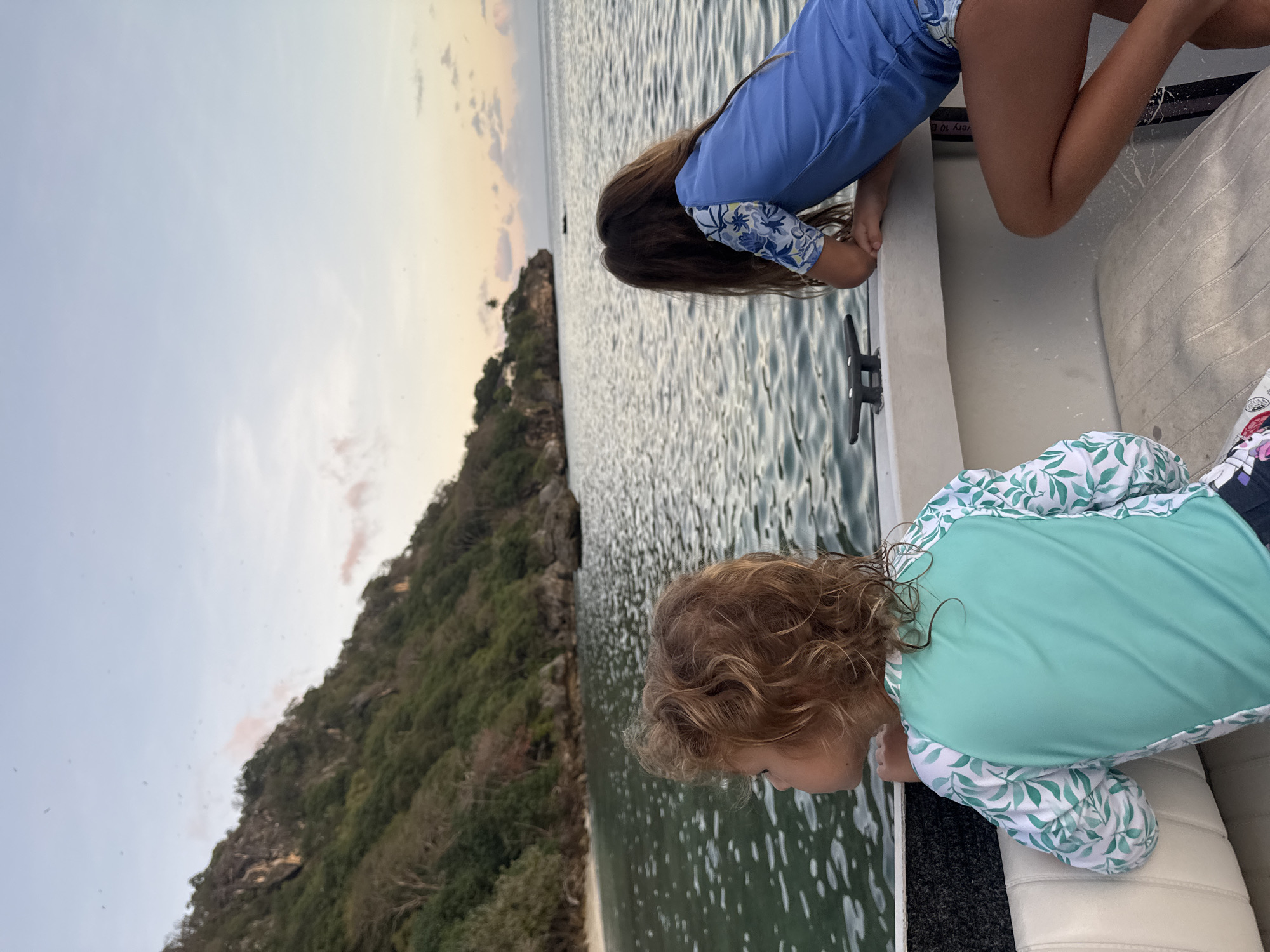 Two young girls are sitting on the edge of a boat, looking out at the ocean. The girl in the foreground has curly blonde hair and is wearing a teal rash guard with a leaf pattern. The girl in the background has long brown hair and is wearing a blue rash guard. The boat is moving on calm water. In the distance, a lush green island with rocky cliffs rises from the sea. The sky above is a pale blue with hints of pink and orange, suggesting it is either dawn or dusk. There is a sandy beach visible on the island. The overall scene depicts a serene moment of observation on the water.