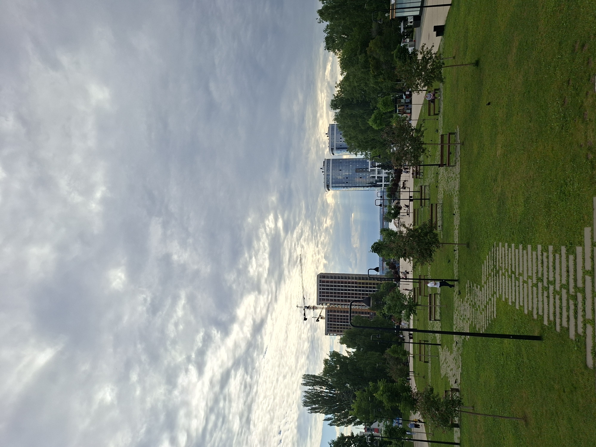 The image shows a park in the city of Izhevsk, Russia, on a cloudy day. In the background, modern buildings and construction cranes are visible. The park features green grass, trees, benches, and pathways. Several people are walking and sitting in the park. The sky is filled with dramatic, gray clouds.