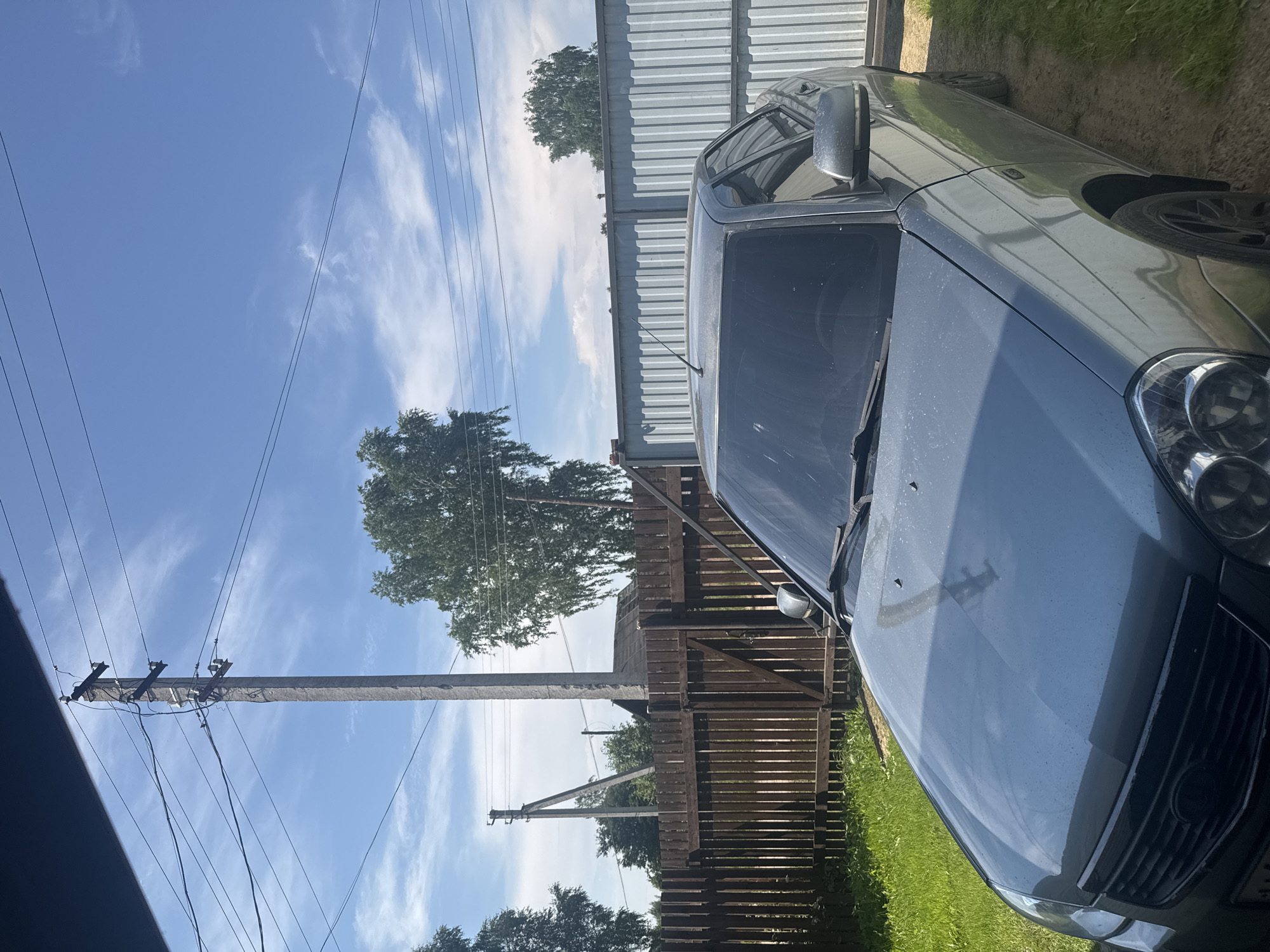 The image shows a gray sedan parked outdoors in the village of Borodulino, Russia. The car is positioned in front of a wooden fence and a corrugated metal fence. A power pole with wires runs vertically in the background, and trees are visible behind the fences. The sky is blue with scattered clouds. The ground is a mix of grass and dirt. No people are visible in the image.