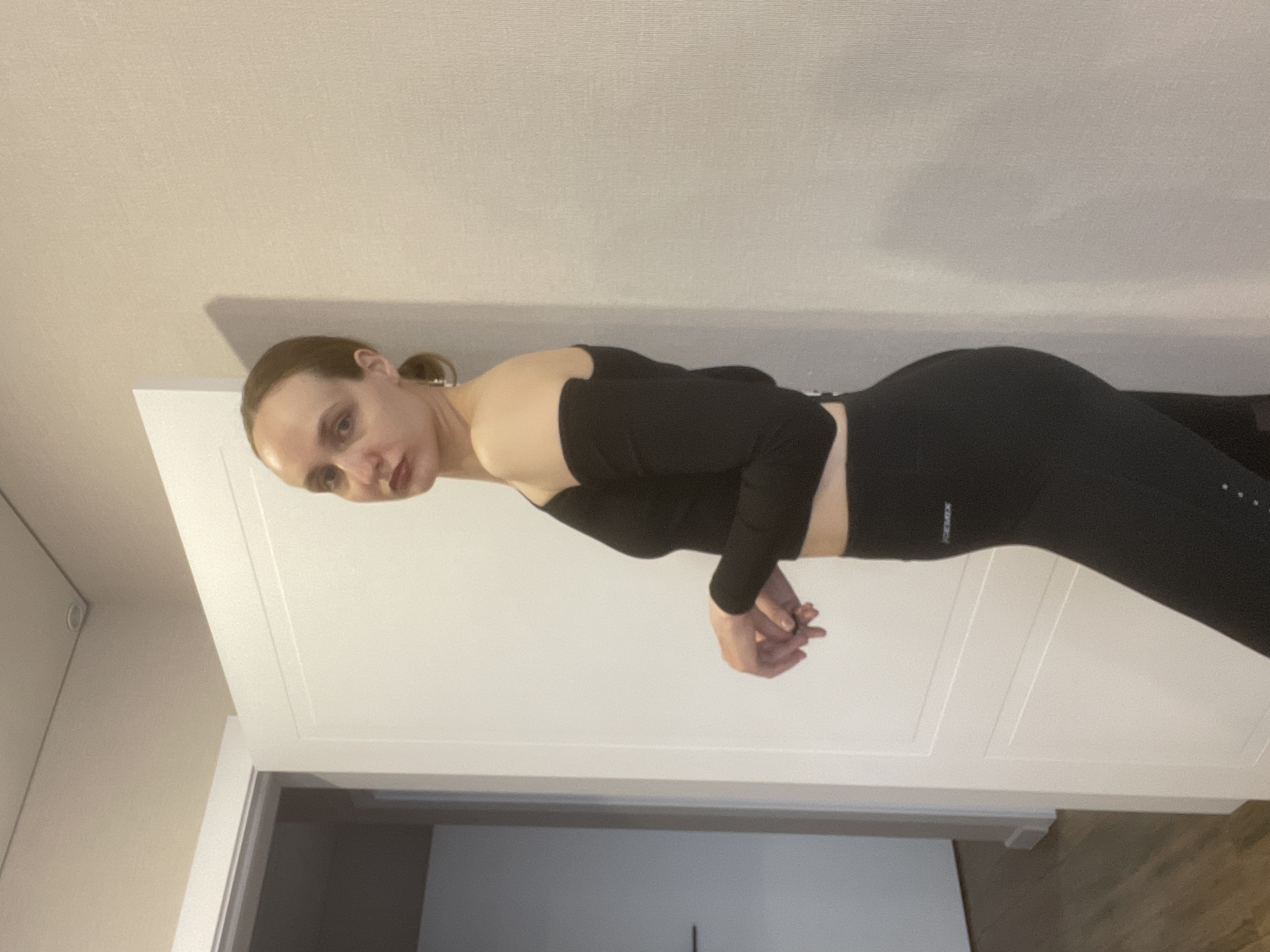 This image captures a full-body shot of a person, likely female, standing indoors in what appears to be a room in Podshivalovo, Russia. The individual has light skin and dark hair tied back in a bun or ponytail. They are dressed in athletic wear, consisting of a black, long-sleeved, off-the-shoulder crop top and matching black leggings. Their hands are clasped together in front of their waist, and they are looking directly towards the camera with a neutral to slightly serious expression.
The background features light-colored walls or white door panels with a panelled design. A dark doorway or opening is partially visible on the bottom left, and a light-colored floor, possibly wood, is seen at the bottom right. The overall lighting suggests an interior setting, likely well-lit during the day.
Notable details include the text "FRESHEX" visible on the waistband of the leggings, and a vertical line of small, light-colored dots running down the side of the right leg of the leggings. The image is rotated 90 degrees clockwise from its original orientation.