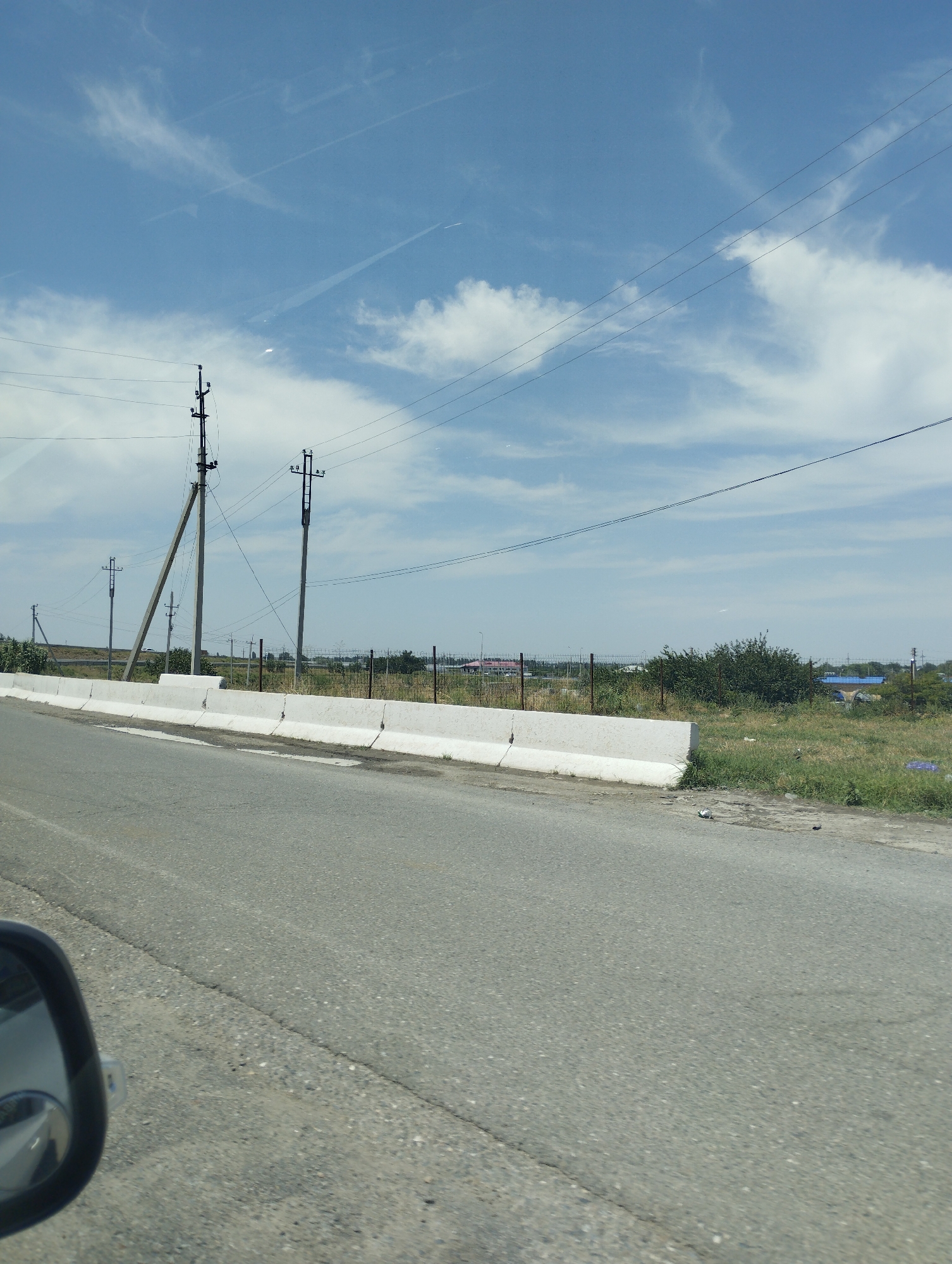 The image is a roadside view in the city of Turkkishlak, Uzbekistan, under a partly cloudy blue sky. In the foreground, an asphalt road is visible, with the side mirror of a vehicle on the left side. A white concrete barrier separates the road from the adjacent area. Behind the barrier, a grassy verge is present, with scattered debris including plastic bags and cans. Further back, a chain-link fence runs horizontally across the frame. Beyond the fence, a line of trees and bushes can be seen, with utility poles and power lines interspersed. In the distance, several low-rise buildings with blue roofs are partially visible, suggesting a rural or semi-urban setting. The overall impression is that of a typical roadside scene in the region.