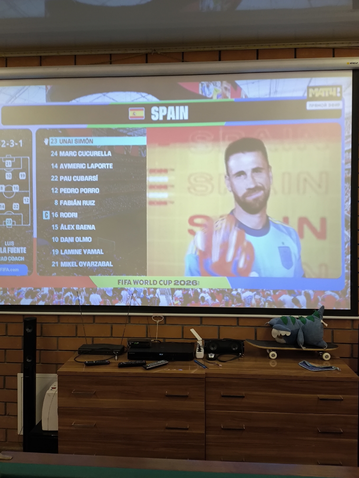 The image shows a projection screen displaying a soccer team's roster for "SPAIN" as part of "FIFA WORLD CUP 2026". On the screen, there is a list of players with their numbers and names, including "23 UNAI SIMON", "24 MARC CUCURELLA", and "14 AYMERIC LAPORTE". There is also a player's portrait on the right side of the screen. Below the screen, a wooden cabinet is visible, on which several electronic devices, including a DVD player and remote controls, are placed. A tall black speaker is positioned to the left of the cabinet. In the background, a brick wall is visible. A stuffed dinosaur toy is sitting on a skateboard on top of the cabinet. The scene appears to be in a domestic or recreational setting, likely in the city of Pokrov, Russia, given the prompt's information.