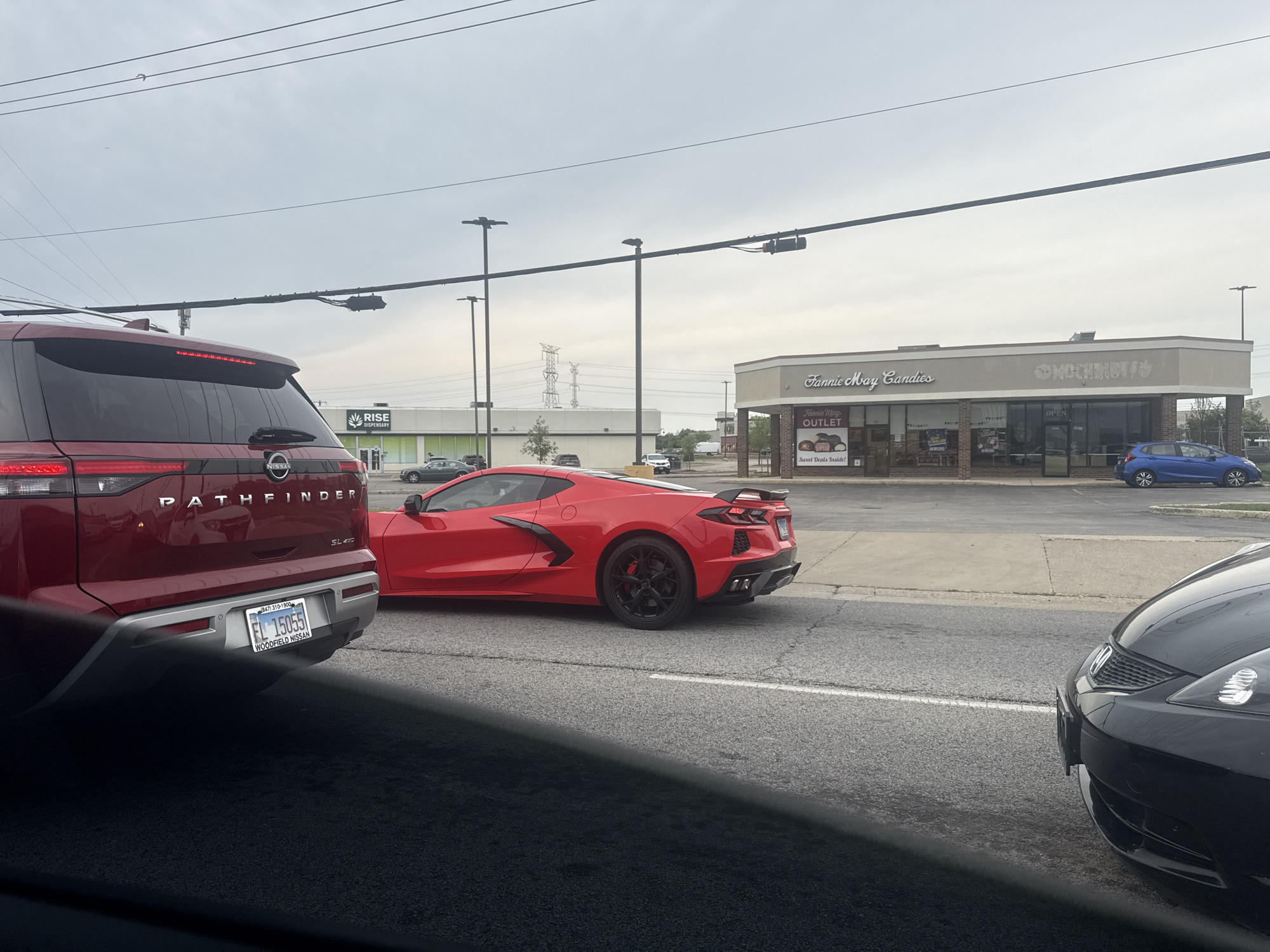 In Niles, Illinois, USA, a red Nissan Pathfinder SUV and a red Chevrolet Corvette are stopped on a road, likely at a traffic light, with other vehicles around. The Pathfinder, in the foreground on the left, has its rear visible, showing a license plate with the text "EL 15055" and "WOODFIELD NISSAN." To the right of the Pathfinder, the red Corvette is positioned, showcasing its sleek design and black wheels.
In the background, a strip mall is visible, featuring businesses such as "Fannie May Candies," an "Outlet," and another establishment with the word "MOCHINUT" visible on its signage. A blue compact car is parked in front of one of the stores. Power lines and utility poles stretch across the sky, and distant industrial structures are visible. The sky is overcast.