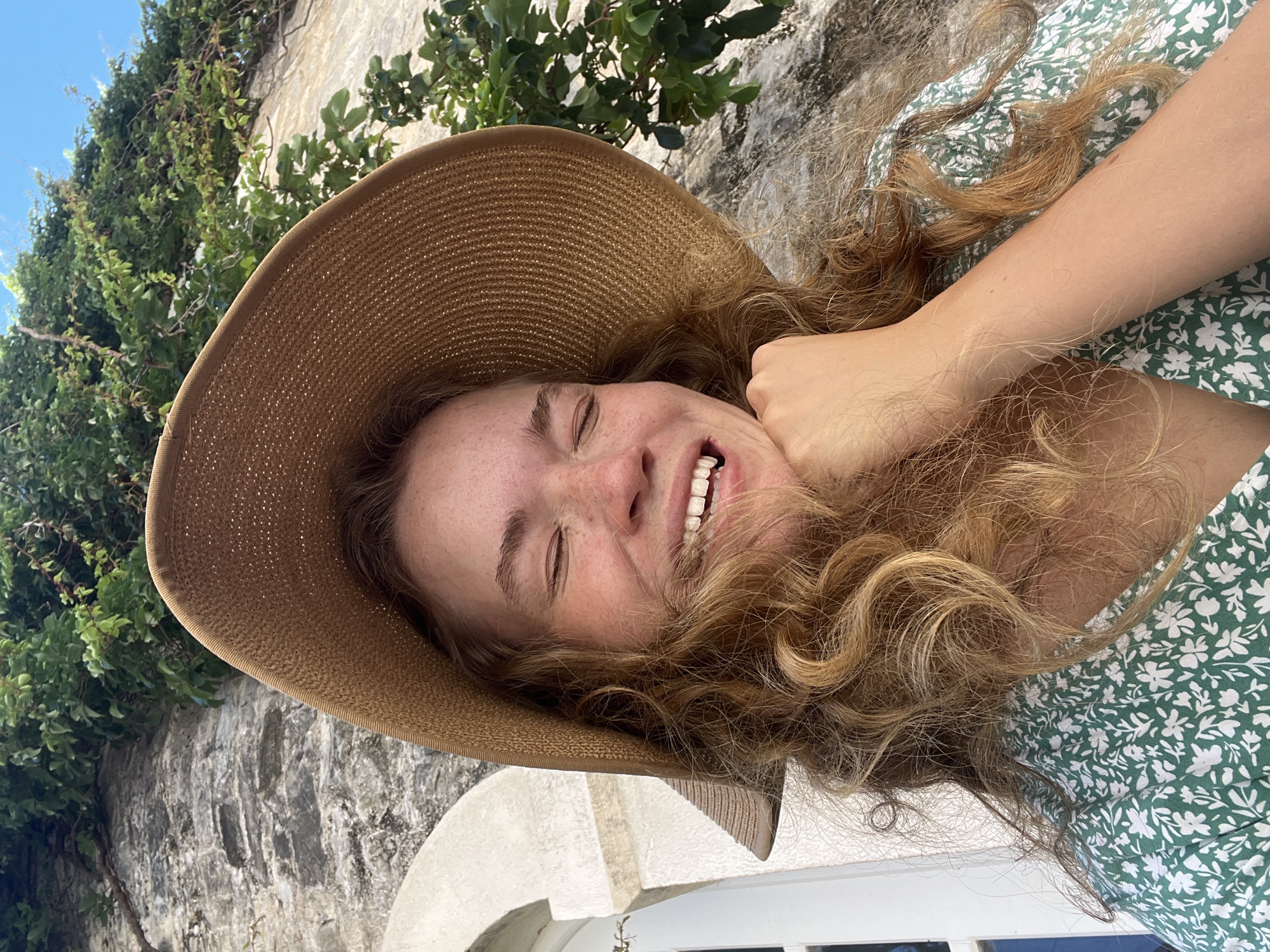 An outdoor, portrait-style photograph captures a young woman from an upward angle, smiling broadly with her eyes squinted or closed. She is wearing a wide-brimmed, woven straw hat of a light brown color and a green dress with a white floral pattern. Her long, wavy, reddish-blonde hair tumbles around her face and over her shoulders, some strands resting on her dress. Freckles are visible across her nose and cheeks, and her white teeth show in her joyful expression. Her right arm, with fair skin and fine hair, is seen resting near her face and neck.
The background features a textured stone wall, partially covered by lush, green leafy vines or ivy, suggesting a natural and old architectural setting. A clear blue sky is visible in the upper left corner. A white arched structure or building element is partially visible at the bottom of the frame. The bright natural light indicates it is daytime, likely sunny. The woman's joyful expression suggests happiness, possibly reacting to the sun or simply enjoying the moment. The overall scene conveys a pleasant, sunny atmosphere in a scenic, possibly Mediterranean, location consistent with Budva, Montenegro. There is no visible text in the image.