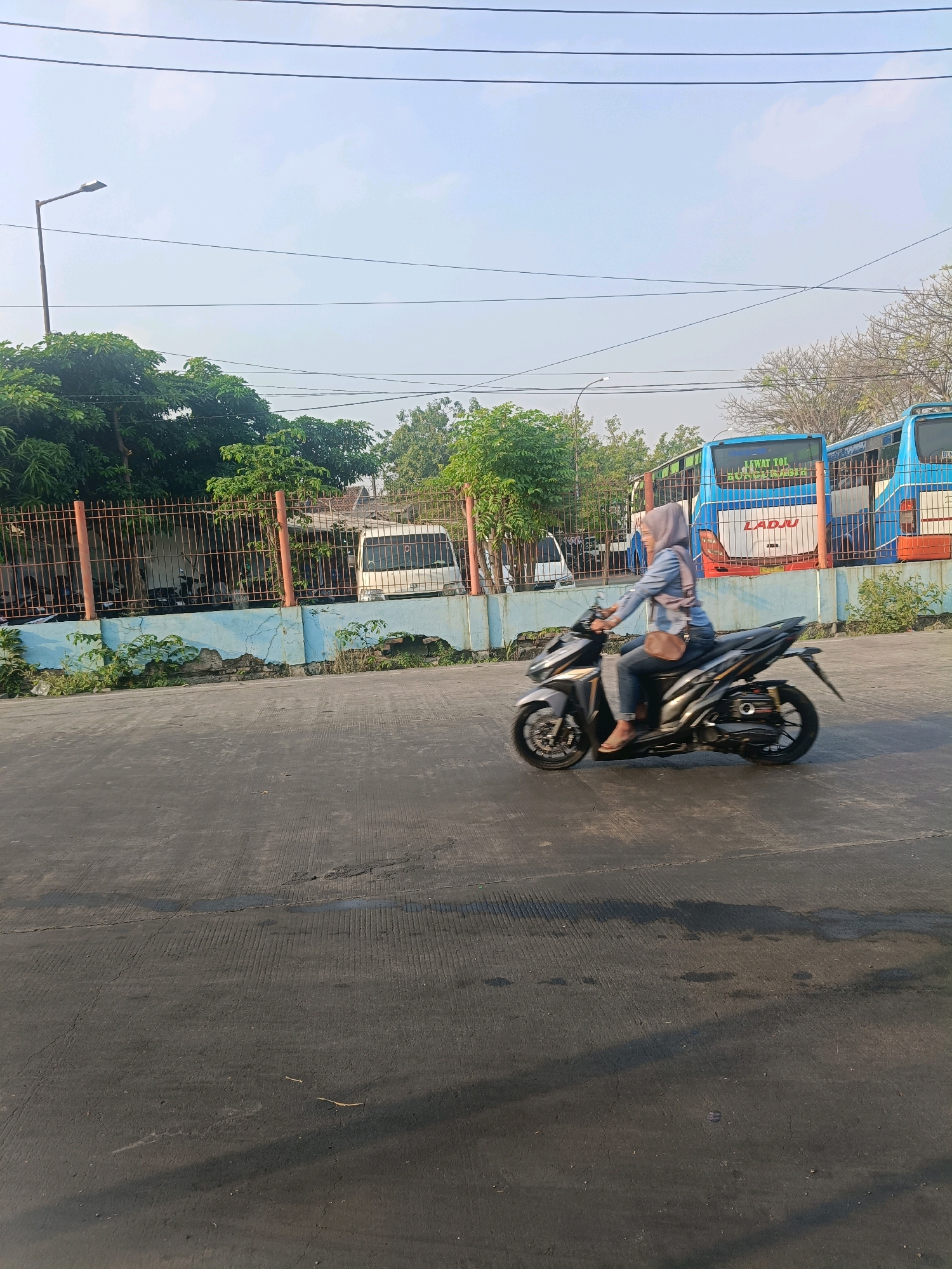 In Kedungrejo, Indonesia, a woman wearing a hijab and a blue denim jacket rides a dark-colored motorcycle on a paved road. She is wearing blue jeans and light-colored shoes. A brown shoulder bag is slung across her body. The motorcycle is in motion, as indicated by the blur of the wheels and the slight tilt of the bike. Behind her, a light blue wall with peeling paint borders a fenced area. Through the fence, several vehicles are visible, including a white van and two blue buses with text that reads "LEWAT TOL" and "LADJU." A street lamp stands to the left of the scene, and utility wires are strung across the sky. The environment appears to be a roadside, possibly near a bus terminal or a public transportation hub. The lighting suggests it is daytime, with the sun casting shadows on the road.