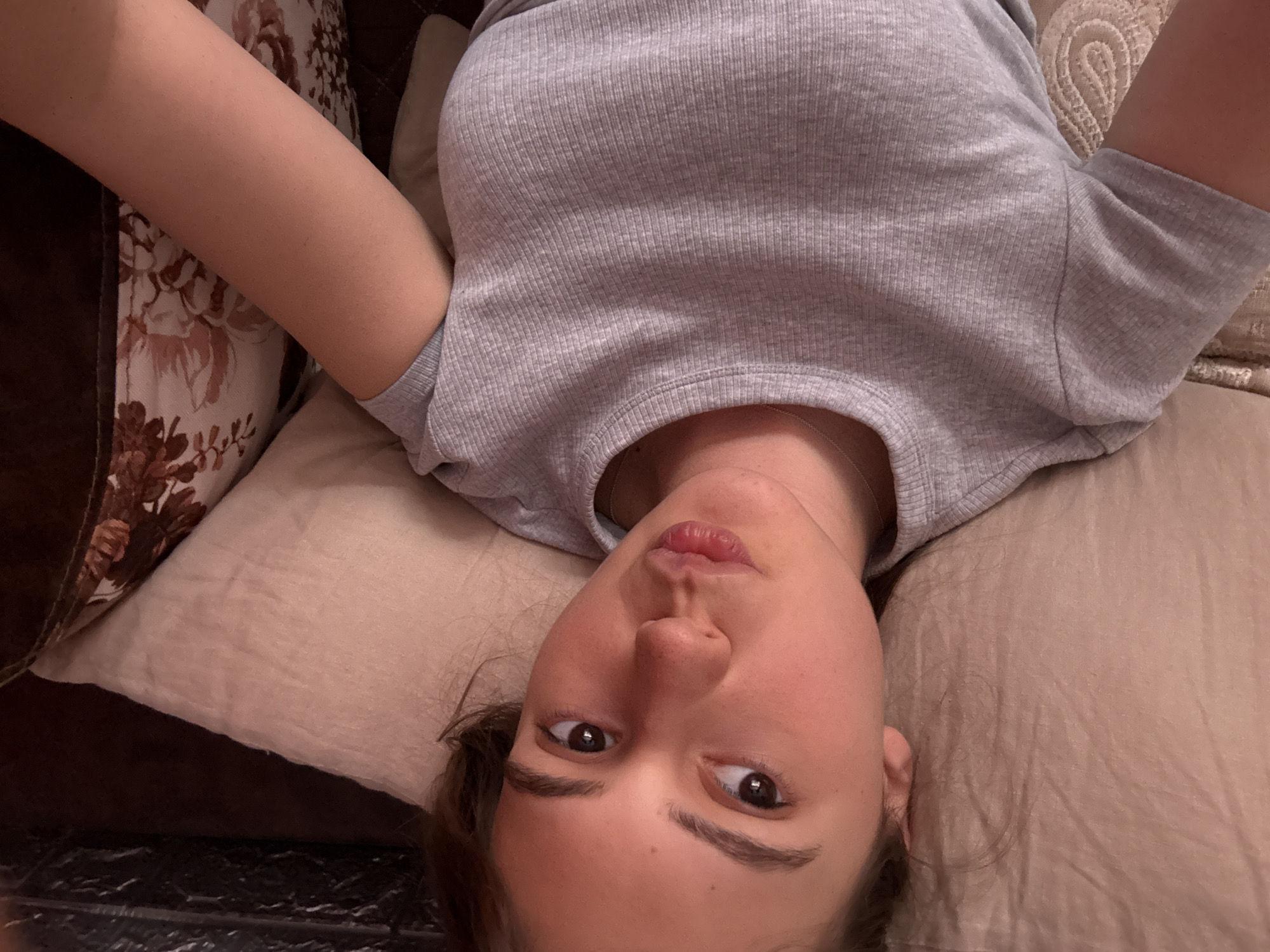 A woman is lying on a pillow, looking directly at the camera. She is wearing a gray ribbed shirt. Her arms are raised. The background includes a floral patterned pillow and a brown cushion. The location is not discernible.