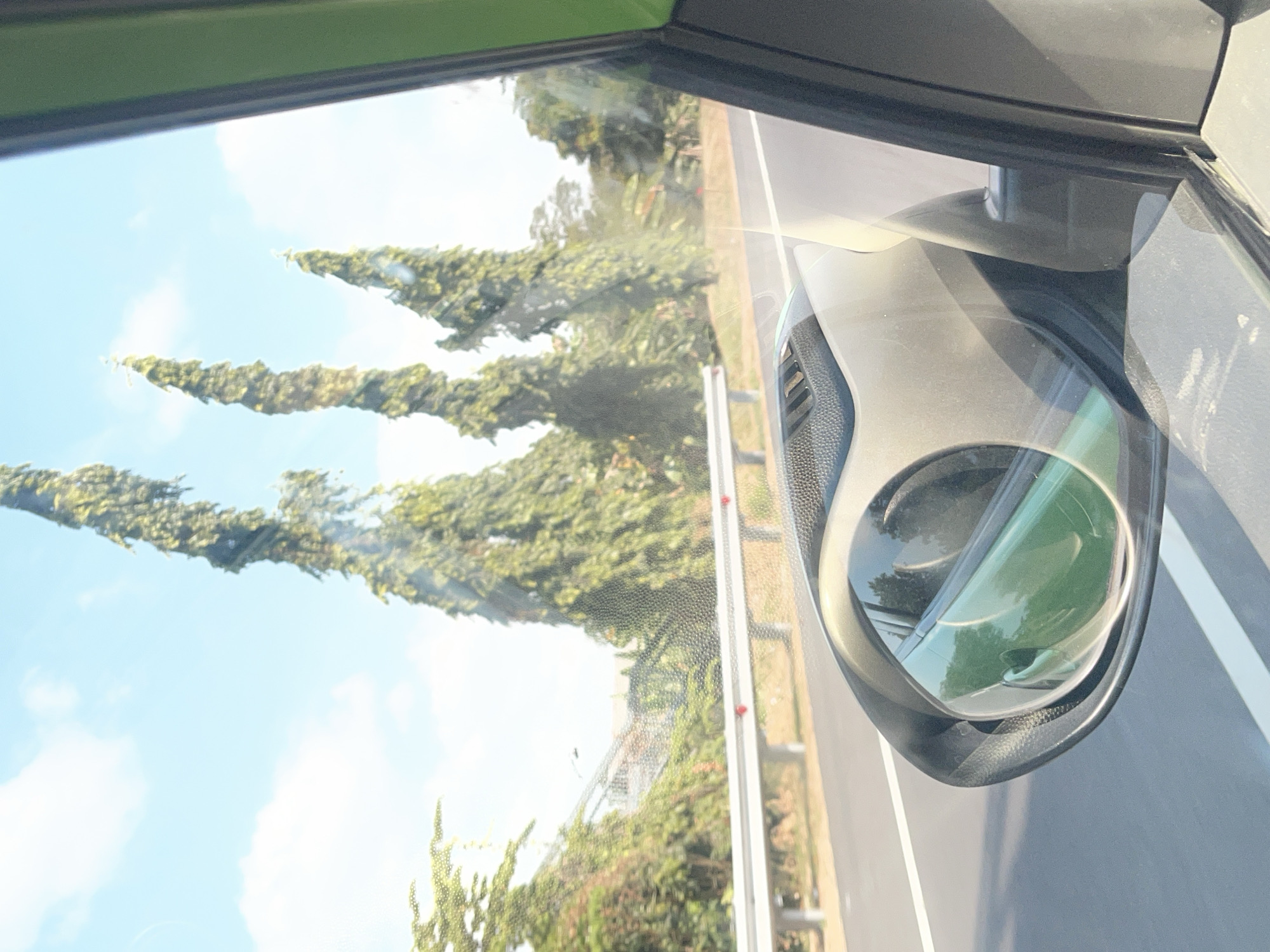 The image is a view from the passenger seat of a car driving on a road, possibly in Kebon Waris, Indonesia, given the context. The perspective is looking forward and slightly to the right, capturing the side mirror and the road ahead. Tall, slender trees with dense green foliage line the right side of the road, with a bright blue sky and scattered white clouds above. A white barrier runs along the side of the road. The car's interior is visible at the bottom and right of the frame, with the green exterior of the car visible on the far right edge. The side mirror reflects the road and the car's interior. The road appears to be paved and has white lane markings. There is no visible presence of people or any animals. The scene depicts a daytime view of a journey on a road surrounded by lush greenery.