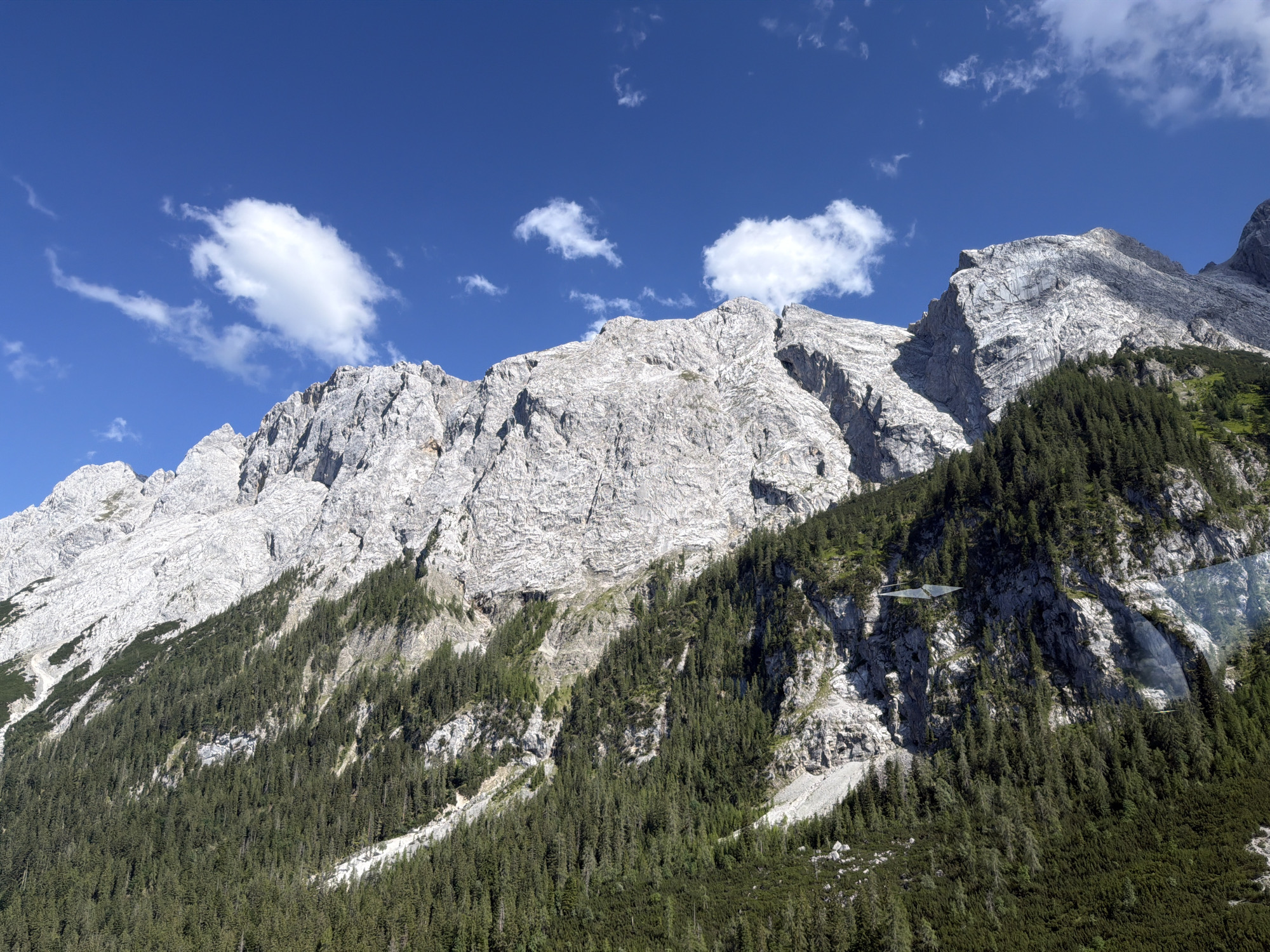 The image depicts a majestic mountain range under a clear blue sky with scattered white clouds. The mountains are rugged and imposing, with sheer rock faces showing signs of erosion and stratification. The lower slopes are covered in a dense forest of dark green coniferous trees, indicating a mountainous, possibly alpine, setting. The sunlight suggests it is daytime, likely midday or early afternoon given the bright illumination.
A notable detail is a light aircraft, possibly a glider or hang glider, suspended in the air against the backdrop of the mountain and trees. Its triangular wings and dark color make it a distinct object against the natural scenery. The overall mood conveyed is one of awe and the grandeur of nature, with the added element of human activity in the form of the aircraft. The location is identified as Grainau, Germany, which is known for its mountainous terrain and outdoor recreational activities. There is no visible text in the image.