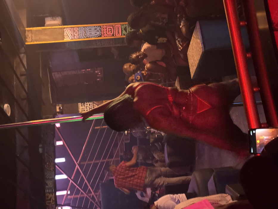 A woman in a red outfit performs on a pole in a crowded club. People are gathered around watching, some with phones in hand. The club has colorful decorative beams with tribal patterns and neon lights. A man in a red plaid shirt leans back, observing the performance. A phone camera captures the scene, with a blurry view of the crowd in the background.