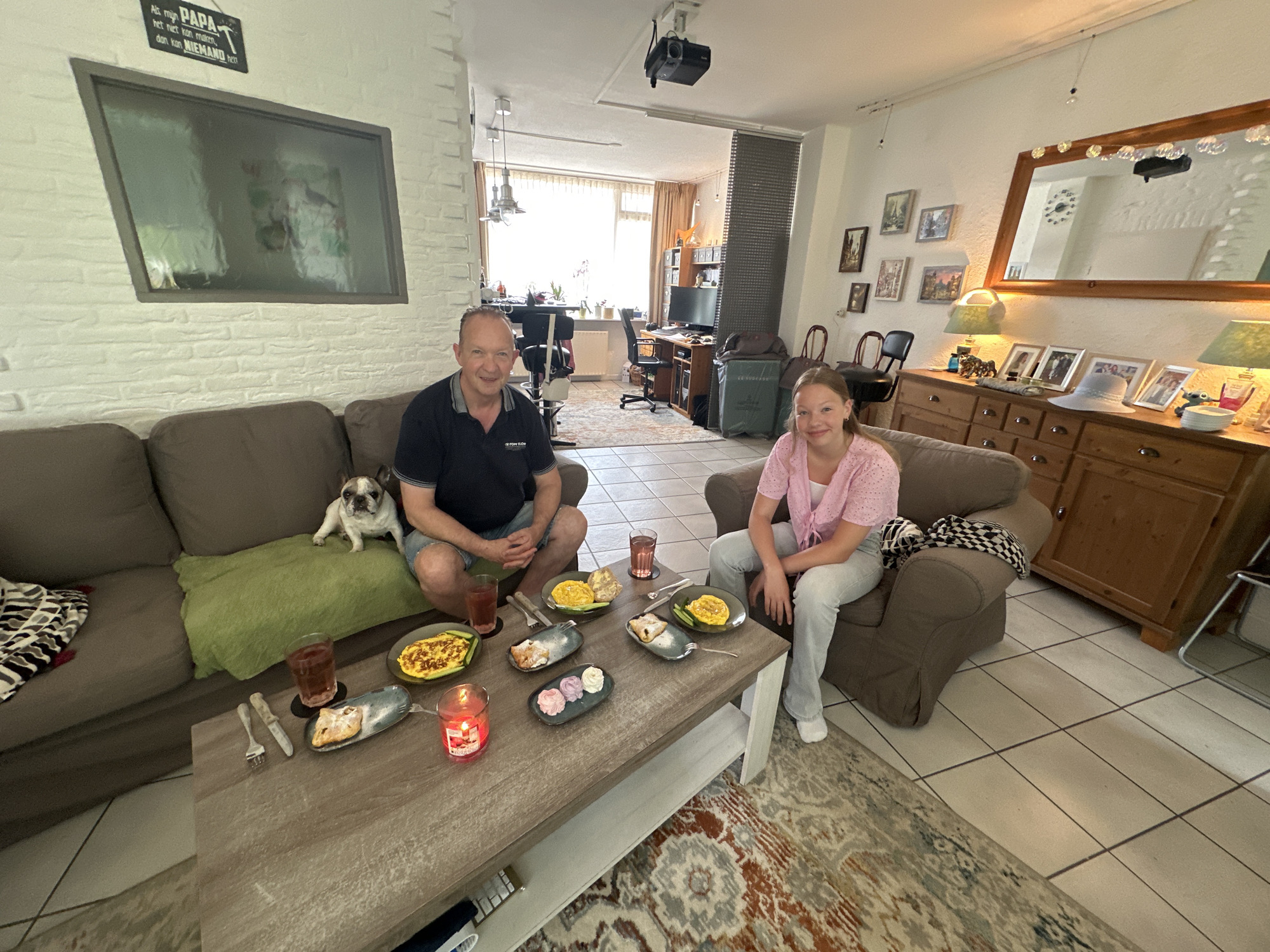 In Steenwijk, Netherlands, a man and a young girl are seated at a coffee table in a living room. The man, wearing a dark blue polo shirt and denim shorts, is on the left side of the frame, with a French bulldog resting on a green cushion beside him. The girl, in a pink top and light blue jeans, is on the right.
The coffee table is laden with food and drinks. Plates of what appears to be omelets are visible, along with a dessert with small pastel-colored mounds and a pastry. Two glasses of red liquid and a lit red candle are also on the table. Utensils, including forks and a knife, are arranged near the plates.
The living room features a dark brown sofa and armchair. In the background, a doorway leads to another room where a desk with a computer and a projector mounted on the ceiling are visible. The walls are painted white, and decorative items such as framed pictures and a large mirror are displayed. A patterned rug covers a portion of the tiled floor.