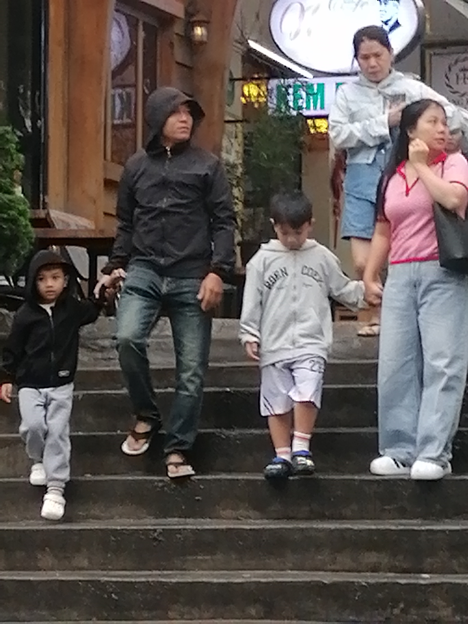 The image captures a family walking down a set of outdoor stone stairs, possibly in an area like a park or a tourist attraction in Da Lat, Vietnam. In the foreground, a young boy in a black hoodie and light grey pants is being led by the hand by an adult male in a black hooded jacket and blue jeans. To the right, a young boy in a grey hooded sweatshirt with "BORN GOOD" printed on it and white shorts walks down the stairs. Beside him, an adult female in a pink t-shirt and light blue jeans is holding his hand. Behind them, another adult female in a light grey hooded jacket and blue shorts stands on the stairs. The background features a wooden building with a rustic design, an outdoor lamp, and some greenery, suggesting a natural or semi-natural environment common in Da Lat. The overall atmosphere appears casual and familial, with the group engaged in the act of descending the stairs.