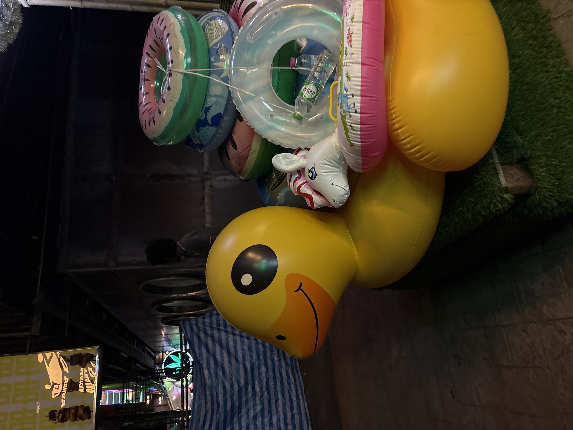 The image shows an outdoor scene at night in Choeng Thale, Thailand, with a focus on inflatable objects. In the foreground, a large, yellow, inflatable duck is prominently featured, facing left. Next to the duck, a stack of inflatable swimming rings is visible, including one designed to look like a watermelon slice, a clear ring with a bottle of water inside, and another ring with a pink border and a cartoonish animal design. The background includes a dark, metallic structure with circular openings, possibly part of a building or stall. Above, a brightly lit sign with Thai text and images of food is partially visible. A person with dark, curly hair can be seen in the middle ground, looking towards the right. The ground appears to be paved with textured tiles in the foreground, transitioning to artificial turf where the inflatables are placed. Blue and white striped tarpaulin is draped over an object to the left. The overall atmosphere suggests a commercial or entertainment area, possibly a street market or a roadside attraction at night.