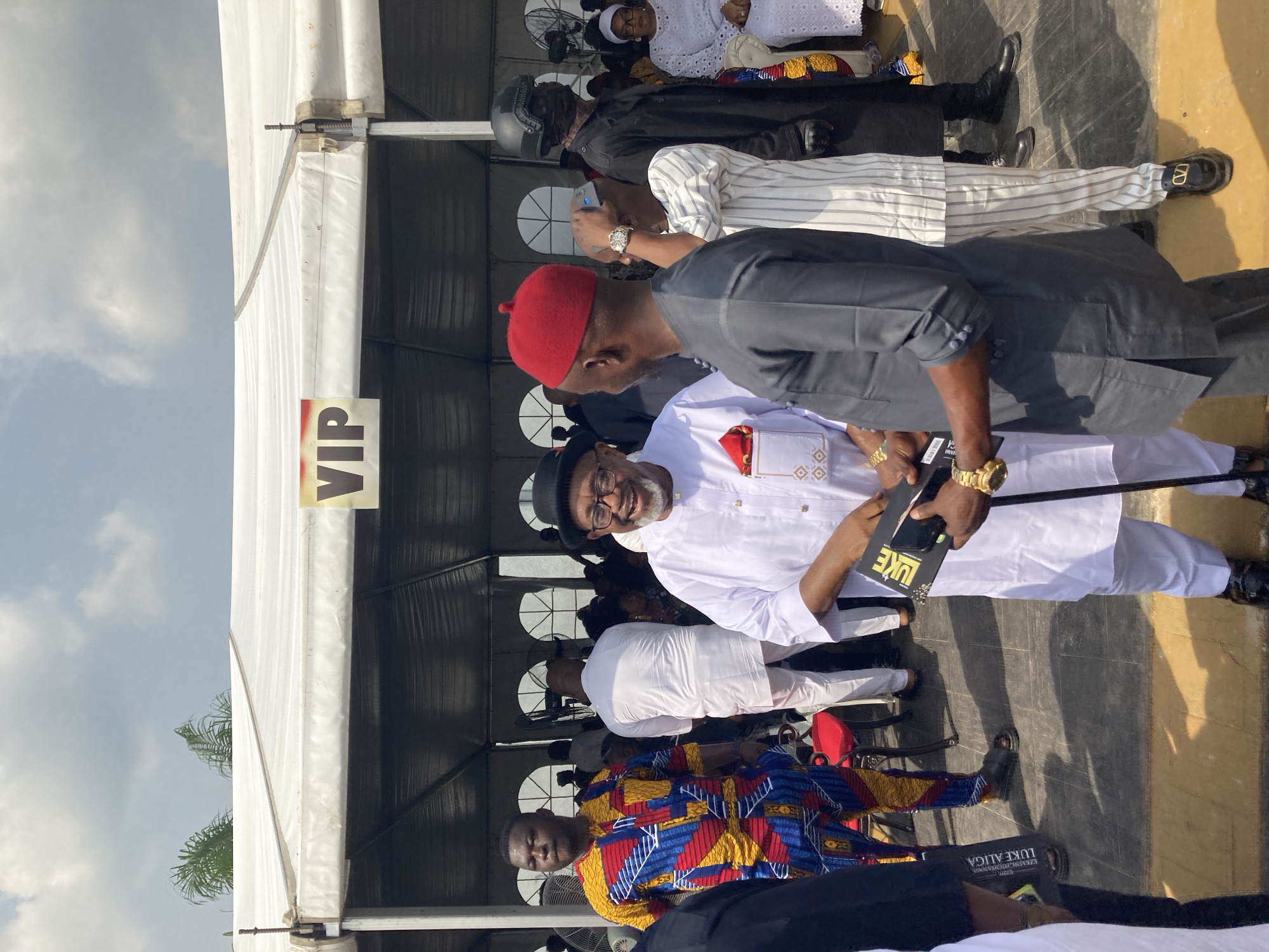 The image shows a group of people at what appears to be an event in Nnenu, Nigeria. A man in a white outfit and a black hat is standing and holding a black object labeled "LUKE." He is interacting with another man wearing a red cap and a dark suit. Other people are seated in the background, some dressed in traditional attire. A tent labeled "VIP" is visible in the background. The setting suggests an outdoor gathering, possibly a formal or celebratory occasion.